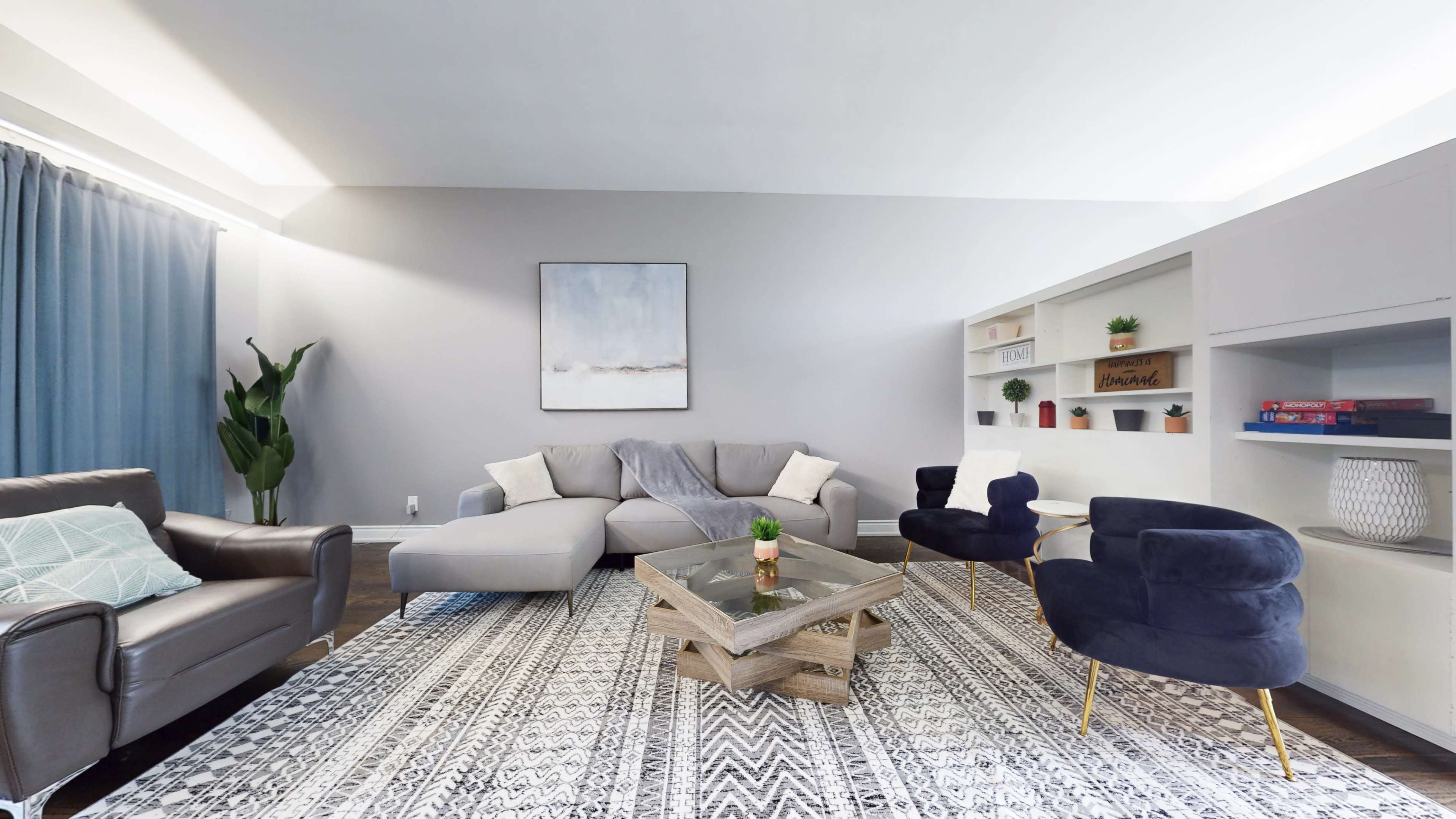 The room features a modern living space with a gray sectional sofa, two dark blue armchairs, a glass coffee table, and a patterned area rug.
