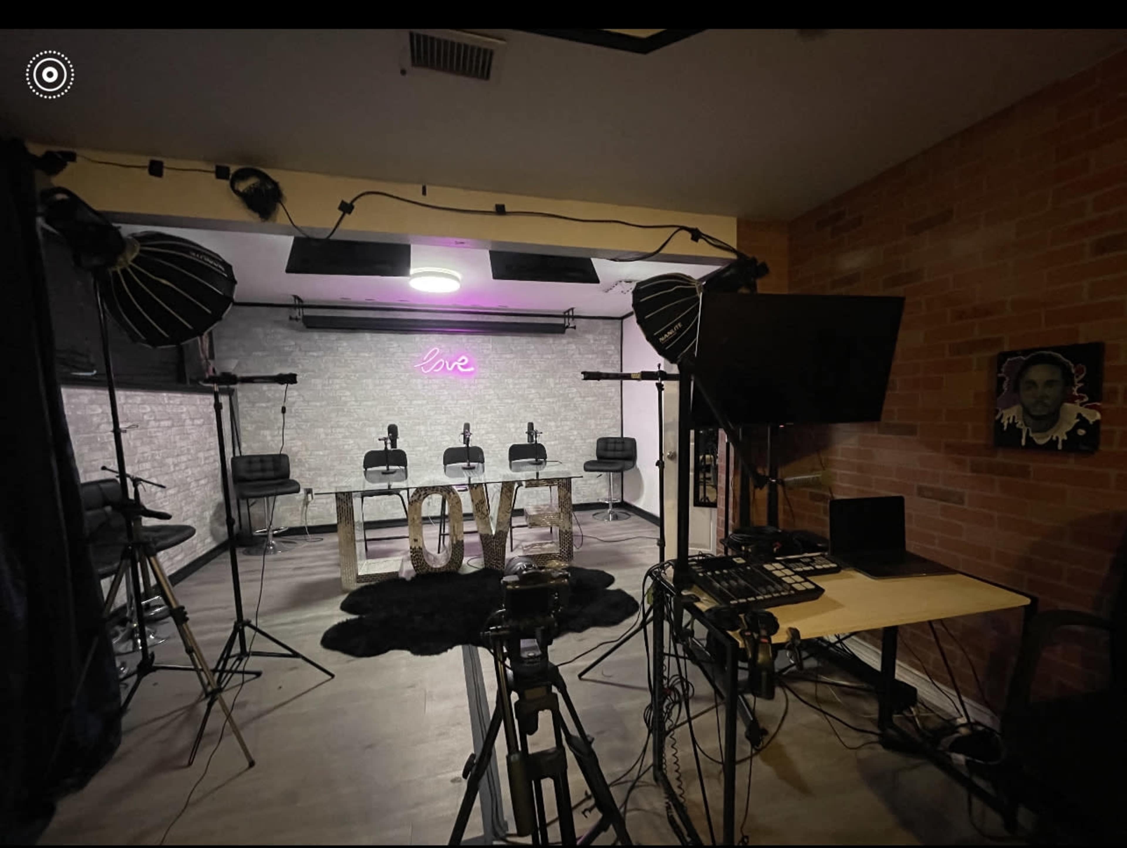 The image shows a studio setup with four chairs facing a lit "live" sign, surrounded by professional lighting equipment and a sound mixer.