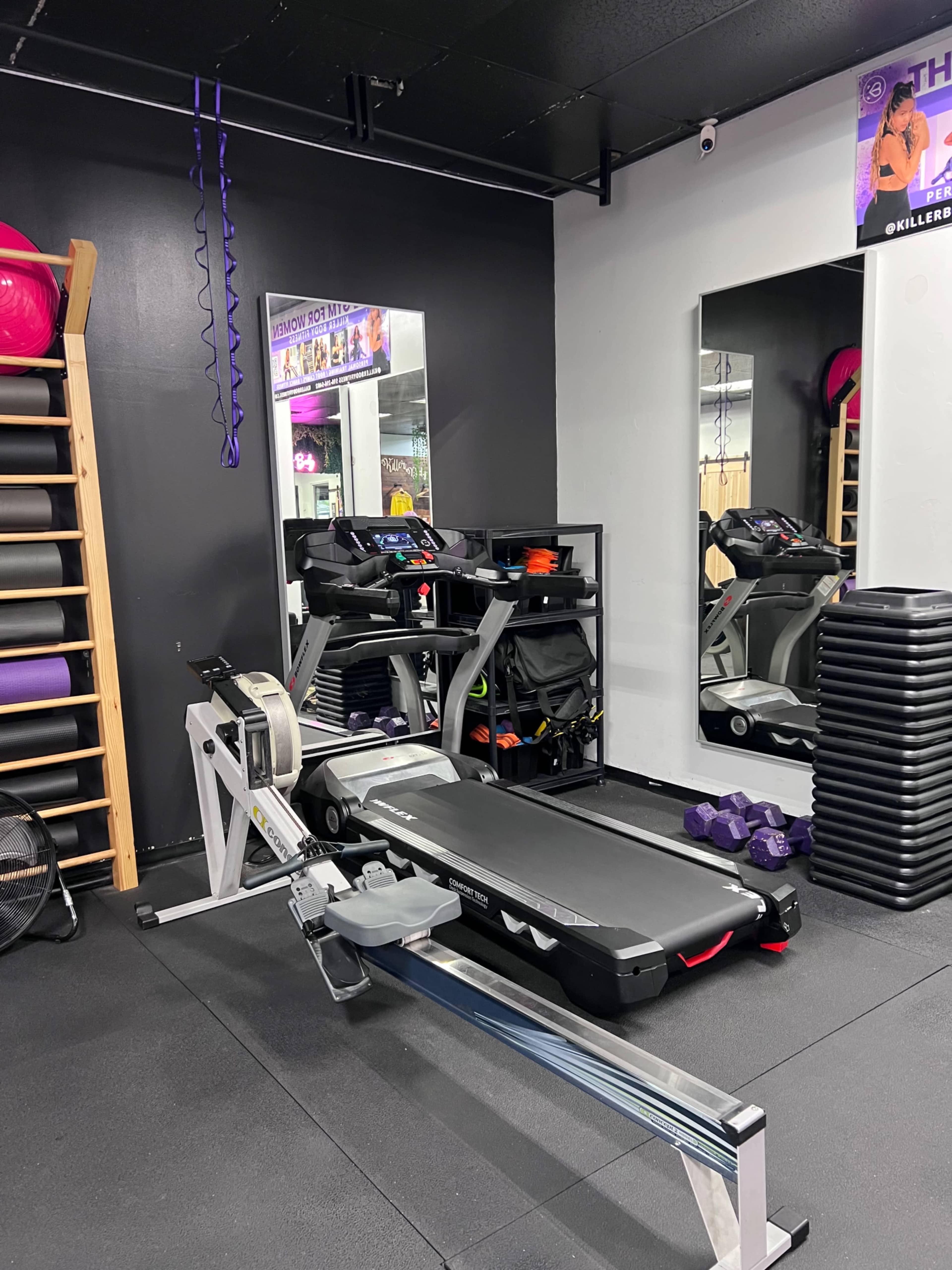 A section of a gym featuring a rowing machine, a treadmill, free weights, and mirrors on a black wall.
