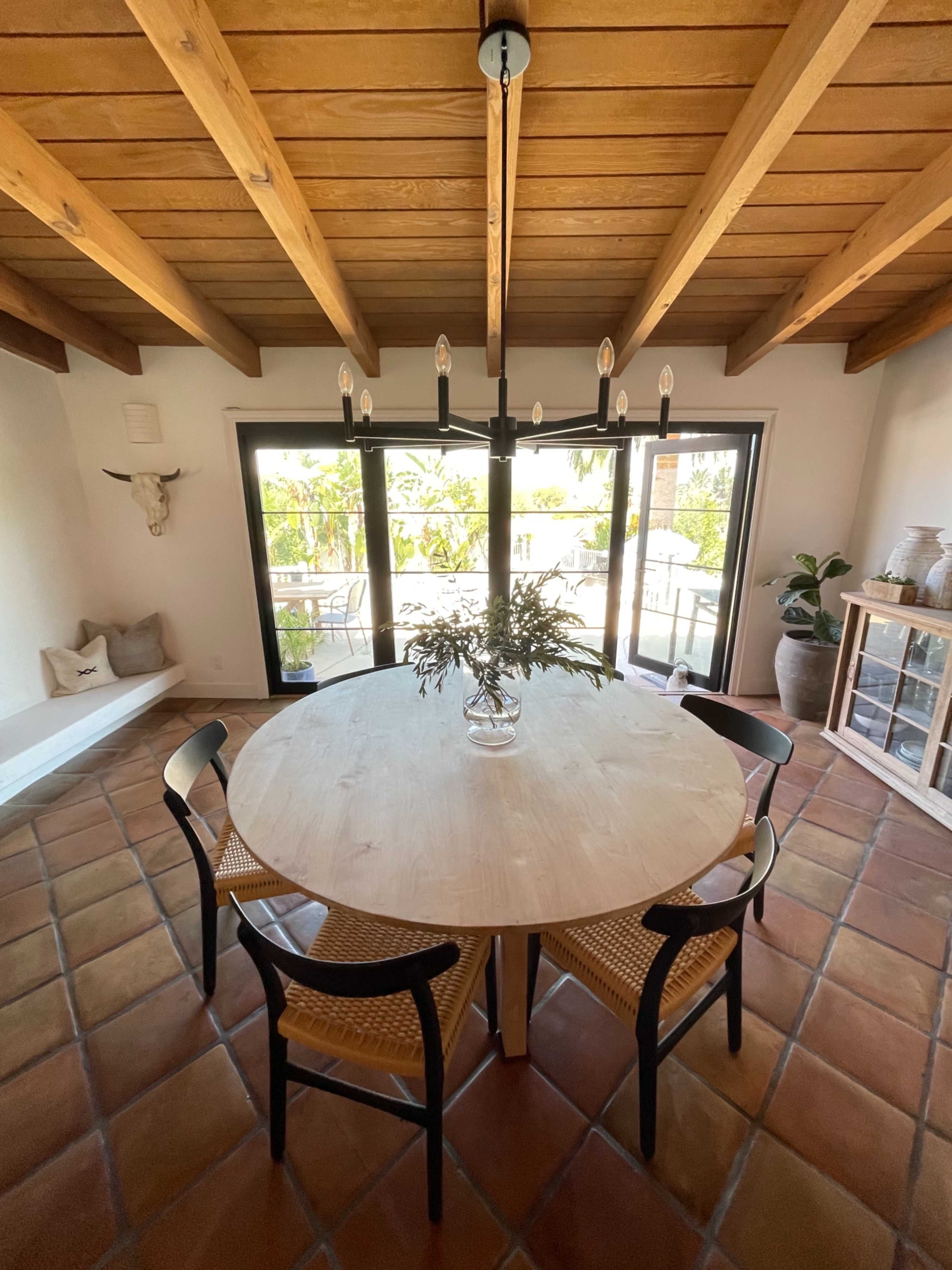 A round wooden dining table with four black chairs is set under a modern chandelier in a well-lit room with sliding glass doors that open to a patio.