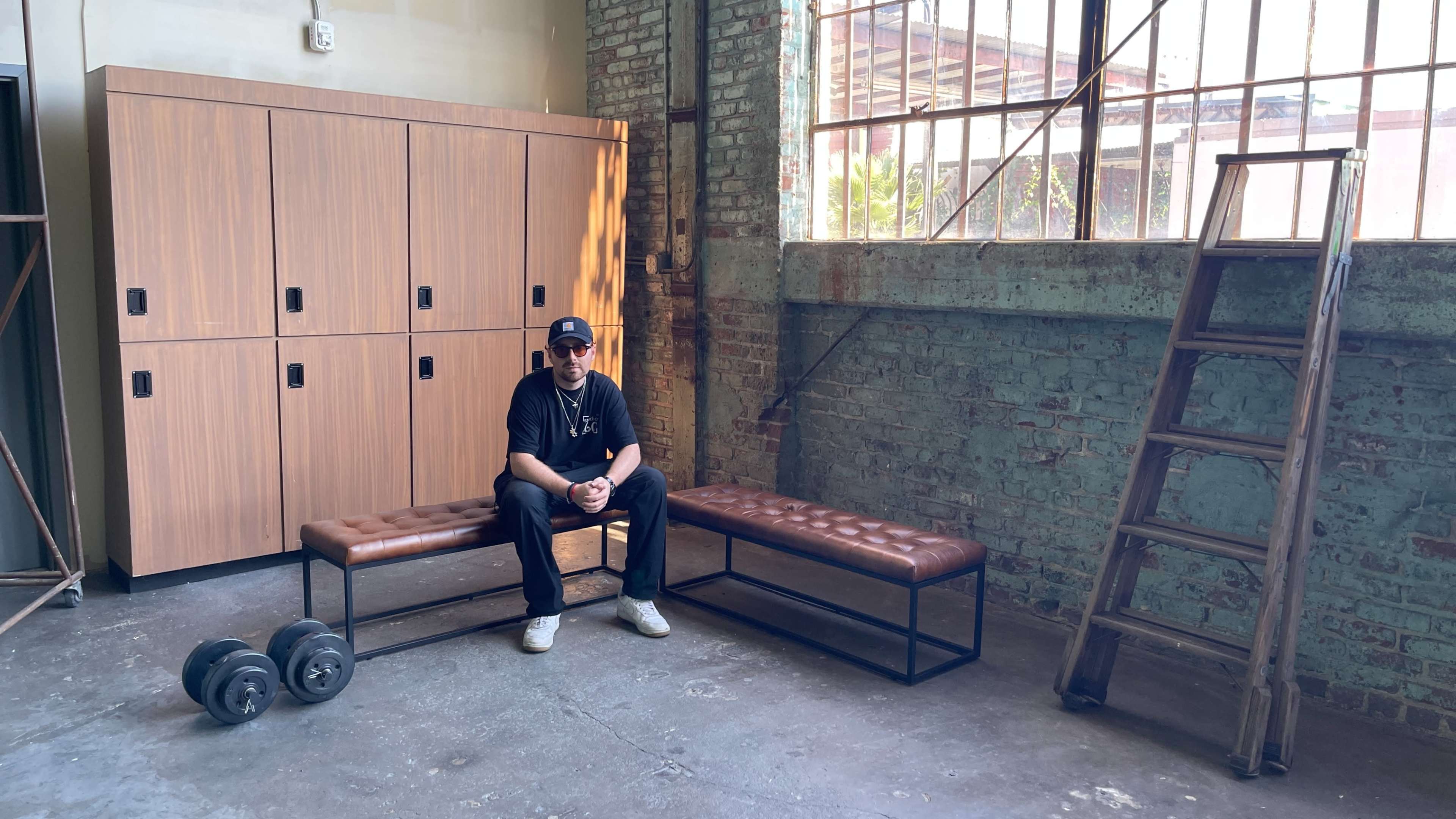 A man sits on a bench in a spacious room with wooden lockers, a metal ladder, and weight plates on the floor.