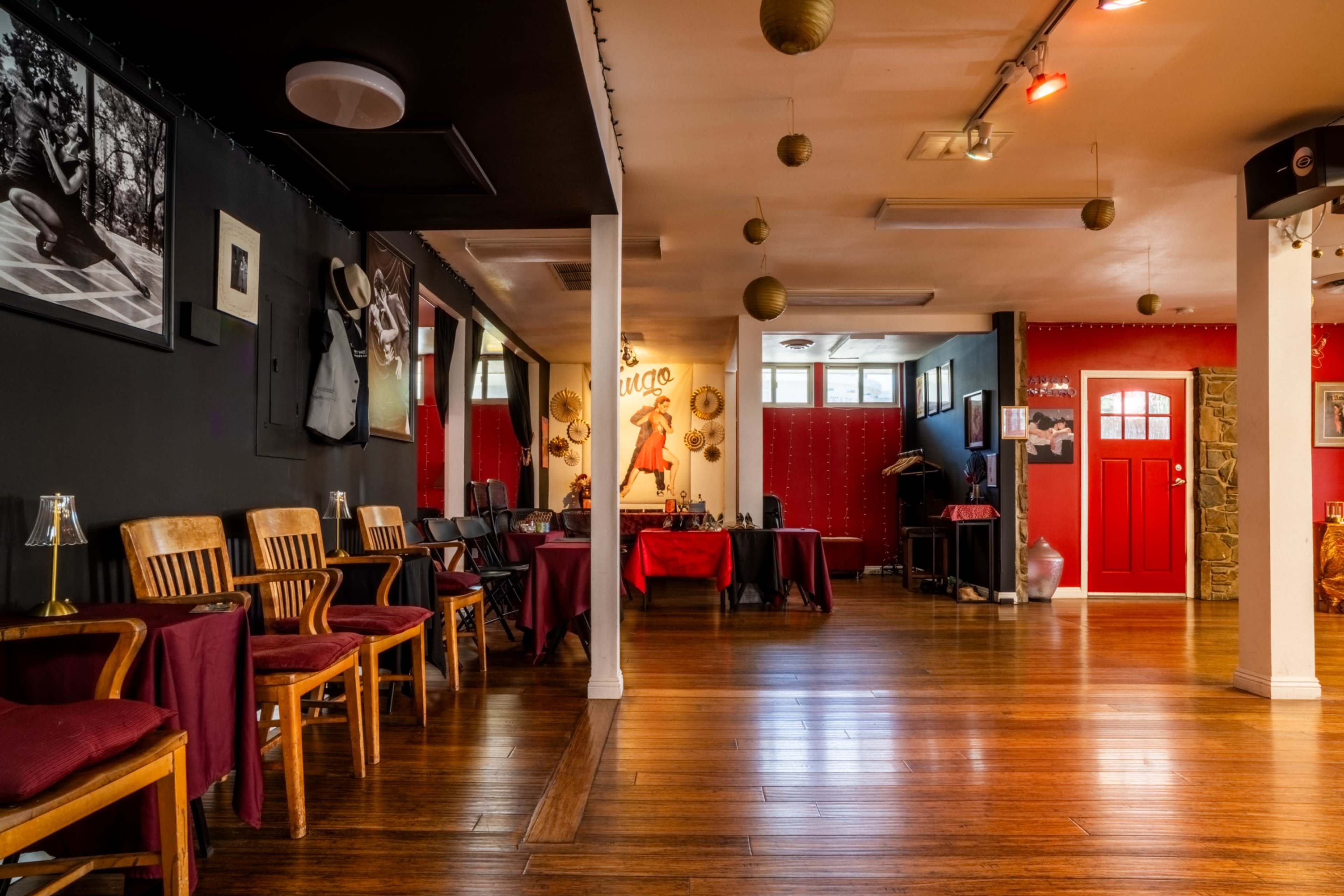 The image shows an interior of a lounge or dance studio with wooden flooring, seating arranged along one side, and a red door leading to another room.