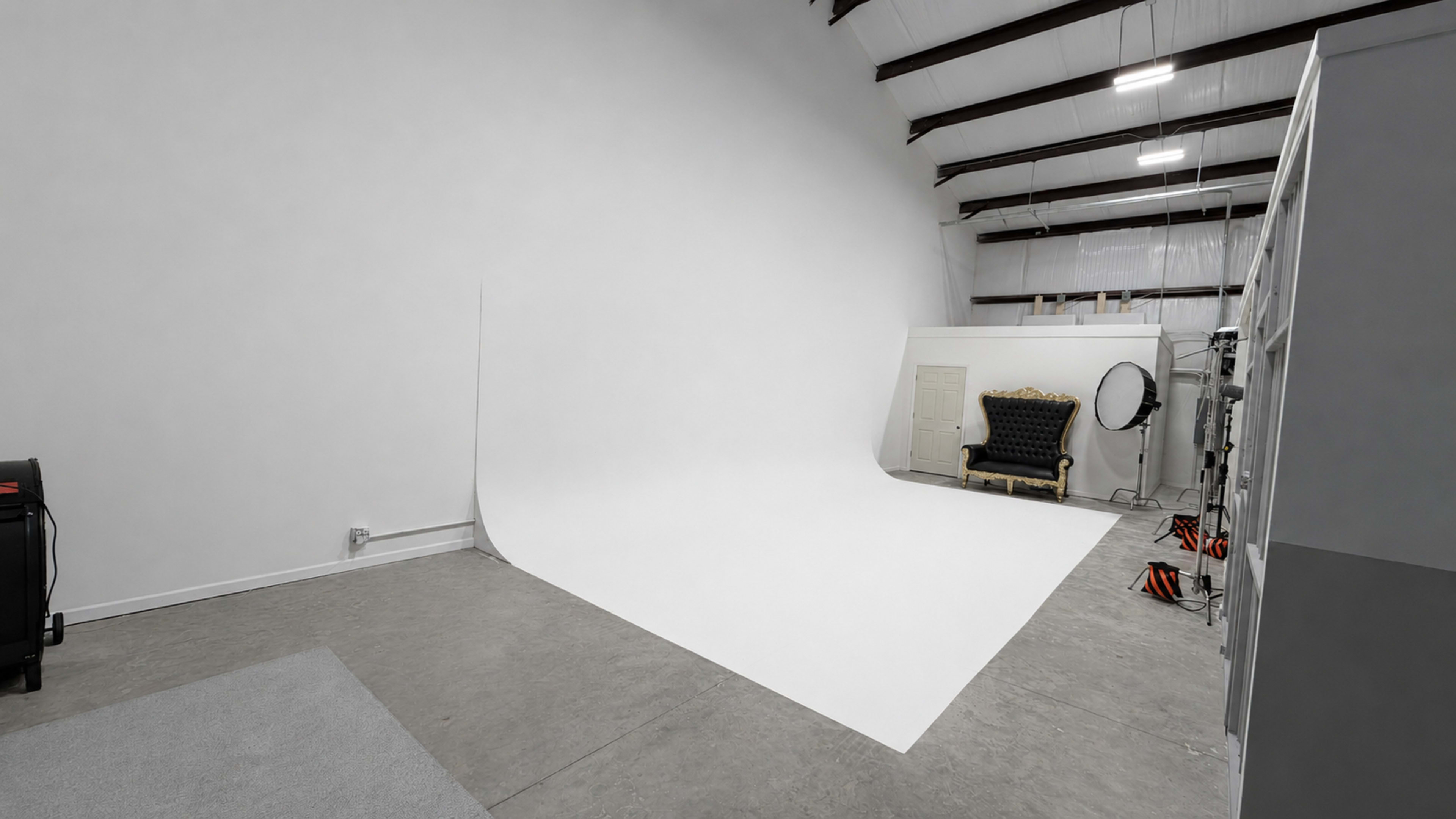 The image shows a spacious photography studio with a large seamless white backdrop, a black ornate chair, and equipment organized along the walls.