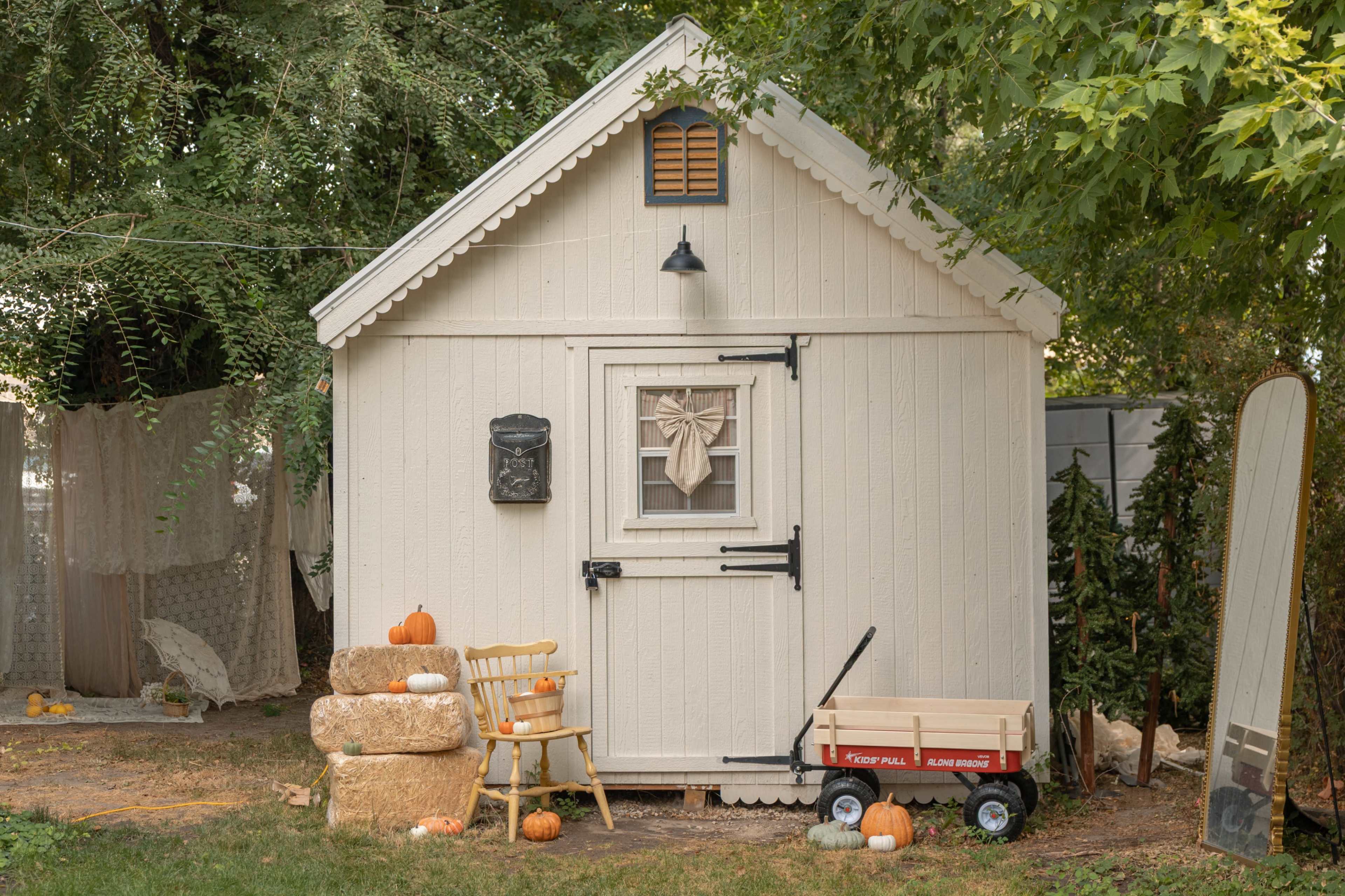 A small white shed is decorated with a wreath and surrounded by pumpkins, hay bales, and a red wagon in a garden setting.