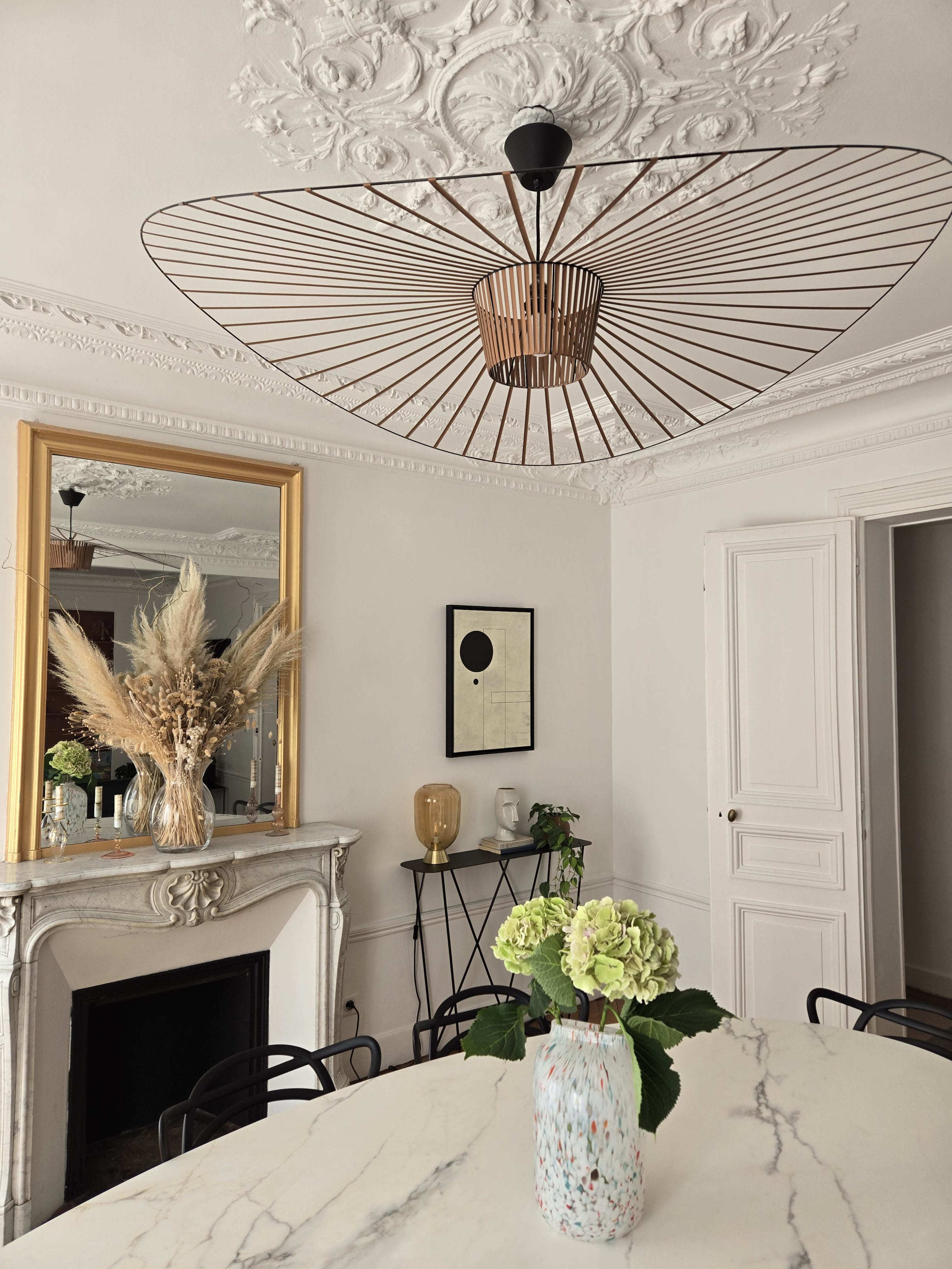 A modern dining area features a large round table with a marble surface, a floral arrangement in a decorative vase, and a striking wire-framed ceiling light above.