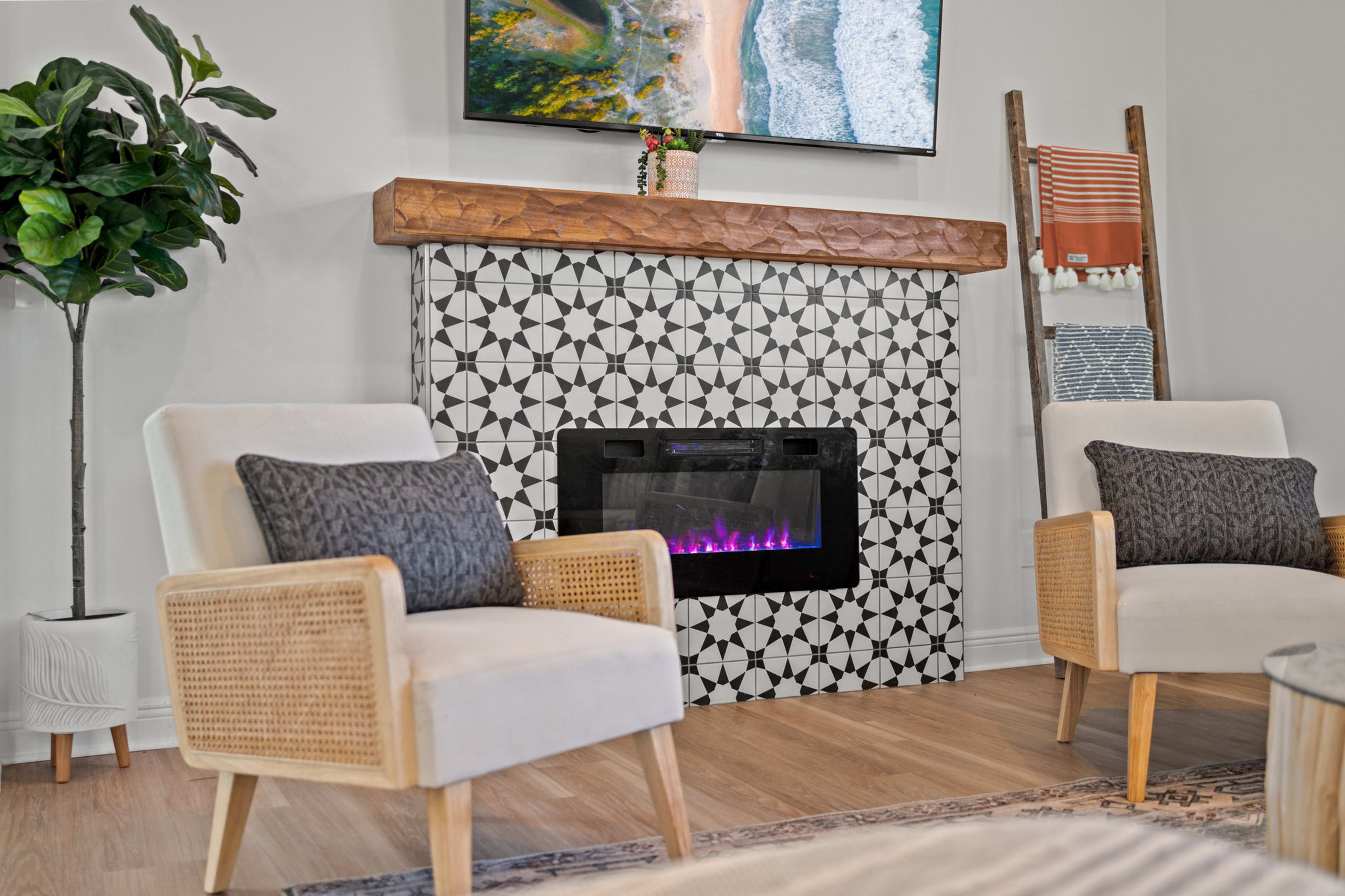 The image shows a modern living room with two armchairs, a decorative tiled fireplace, and a mounted television above a wooden mantel.
