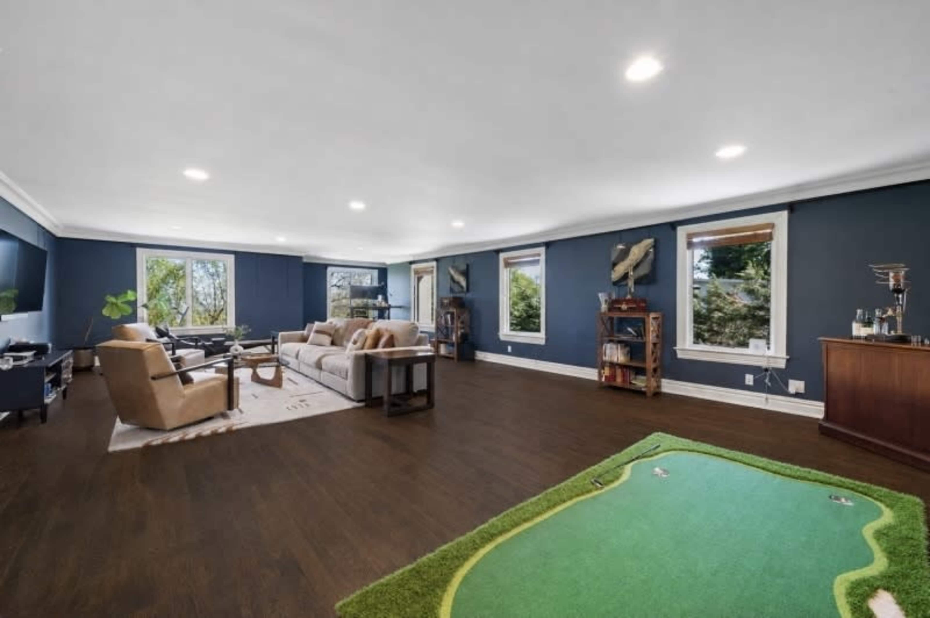 The image shows a spacious living room with dark wood flooring, blue walls, large windows, and a mini putting green in one corner.