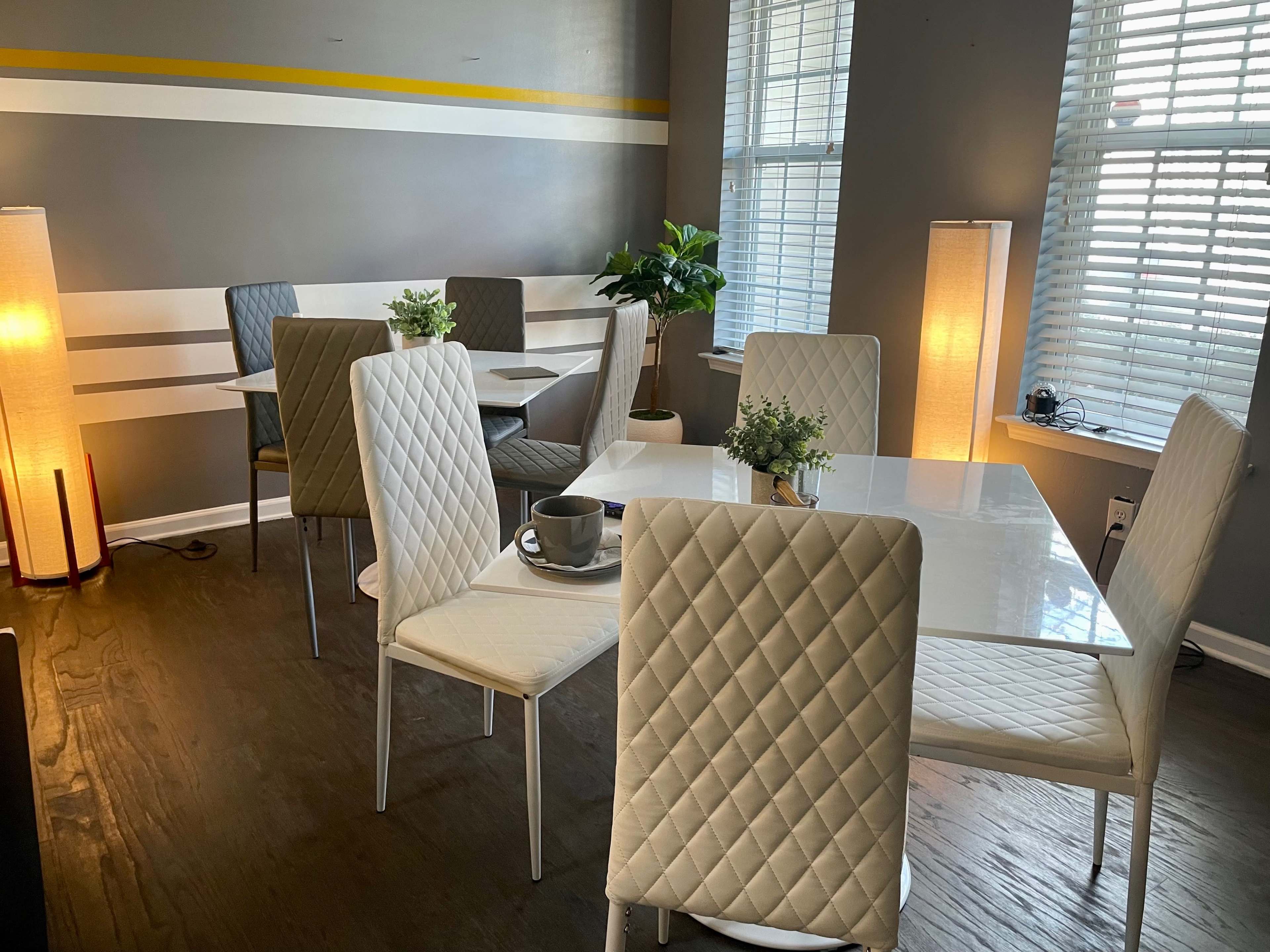 A modern dining area with white and gray furniture, featuring tables and quilted chairs, along with potted plants and ambient lighting.
