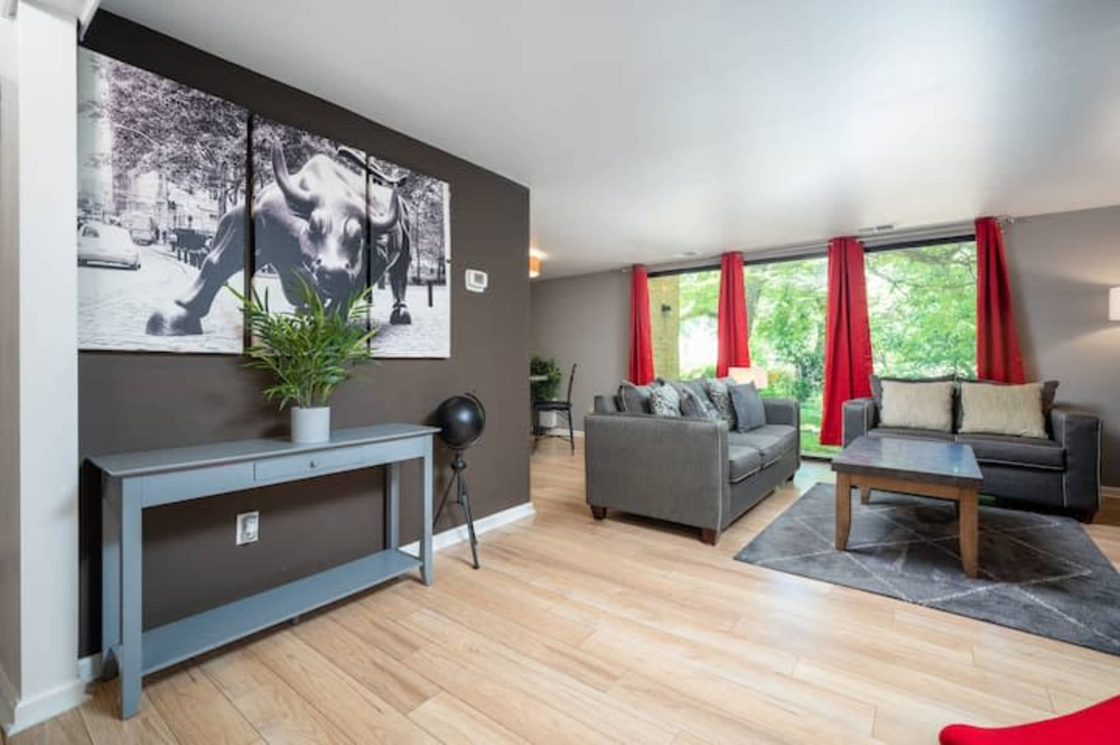The living room features a grey sofa, a dark wooden coffee table, a console table against the wall, and large windows with red curtains allowing natural light to enter.