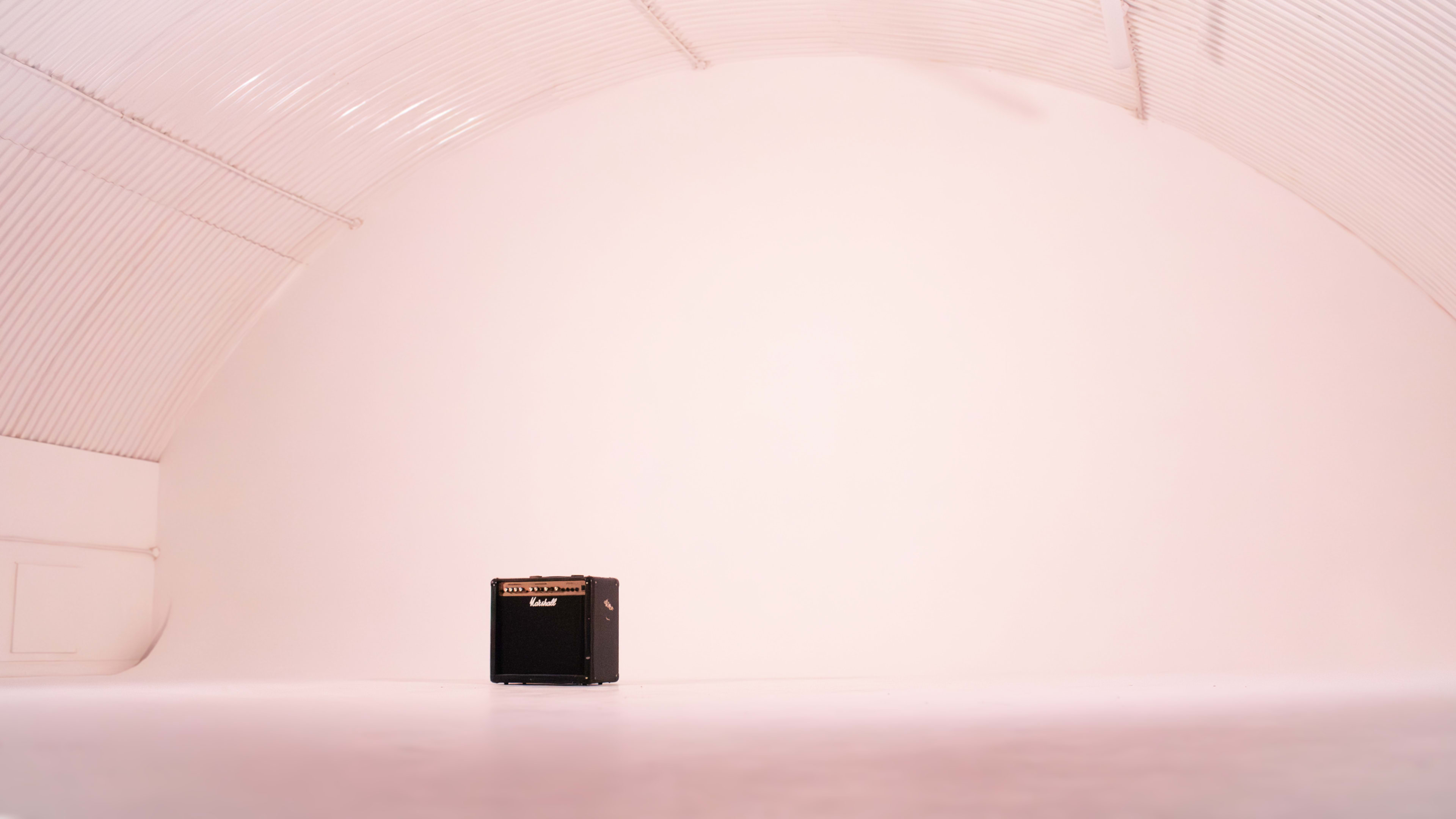 A black guitar amplifier stands alone on a blank white floor within a curved white-walled space.