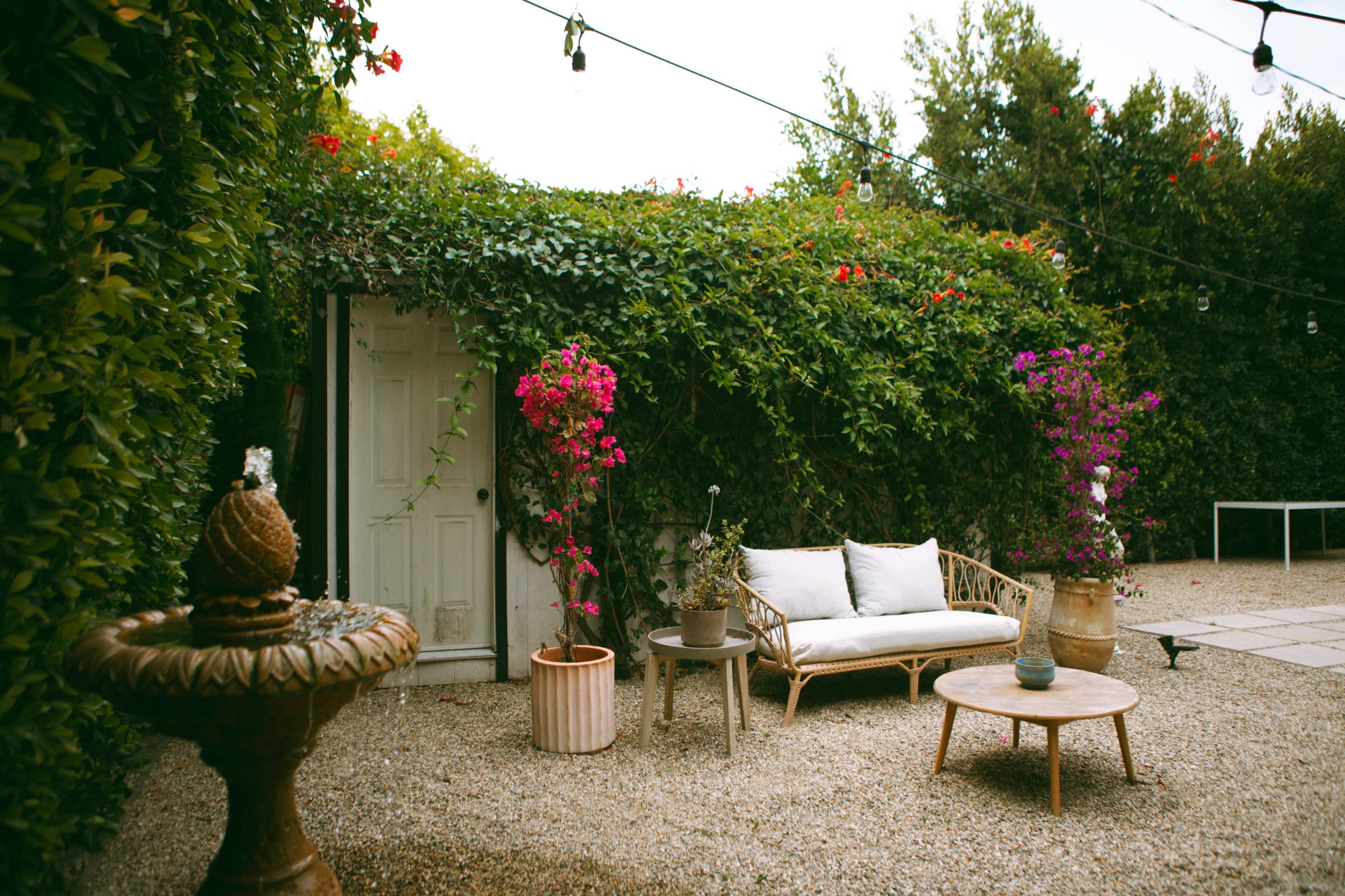The image shows a cozy outdoor seating area surrounded by greenery, featuring a sofa, a fountain, and decorative potted plants.
