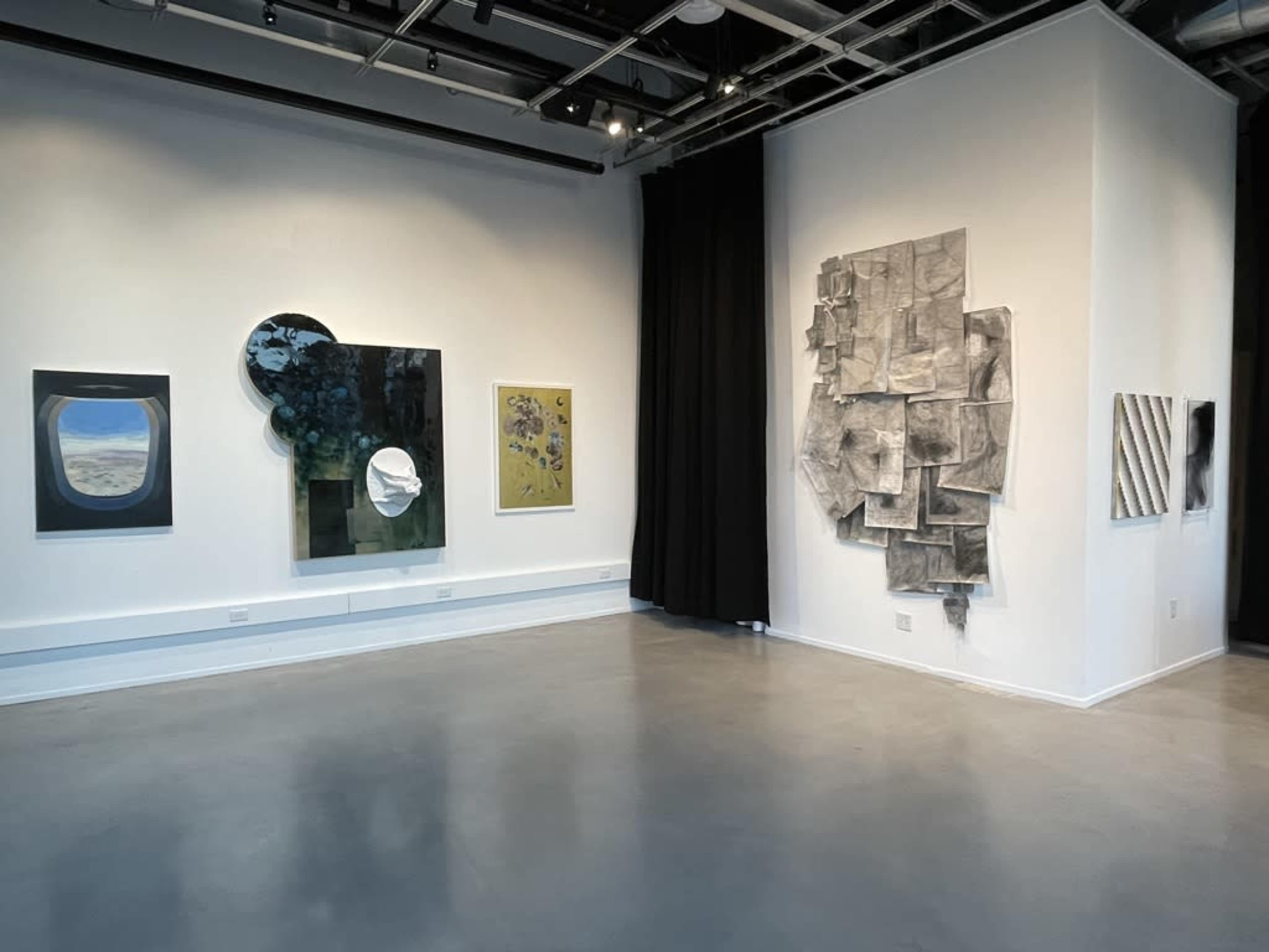 The image shows a gallery interior with several framed artworks displayed on white walls, including abstract pieces and a large textured installation.