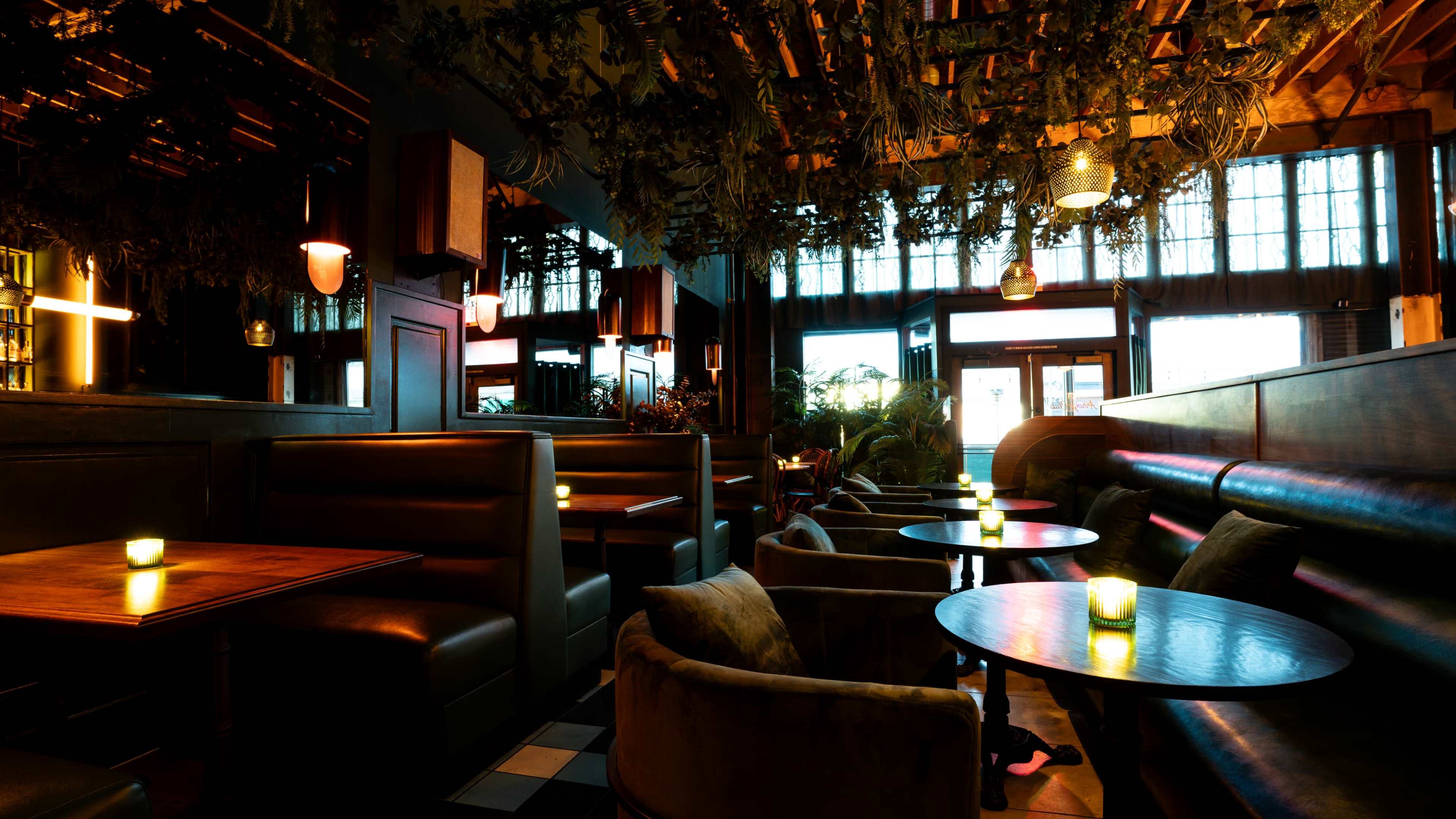 The image shows a modern restaurant interior featuring dark booths, circular tables, and warm lighting, with plants hanging from the ceiling.