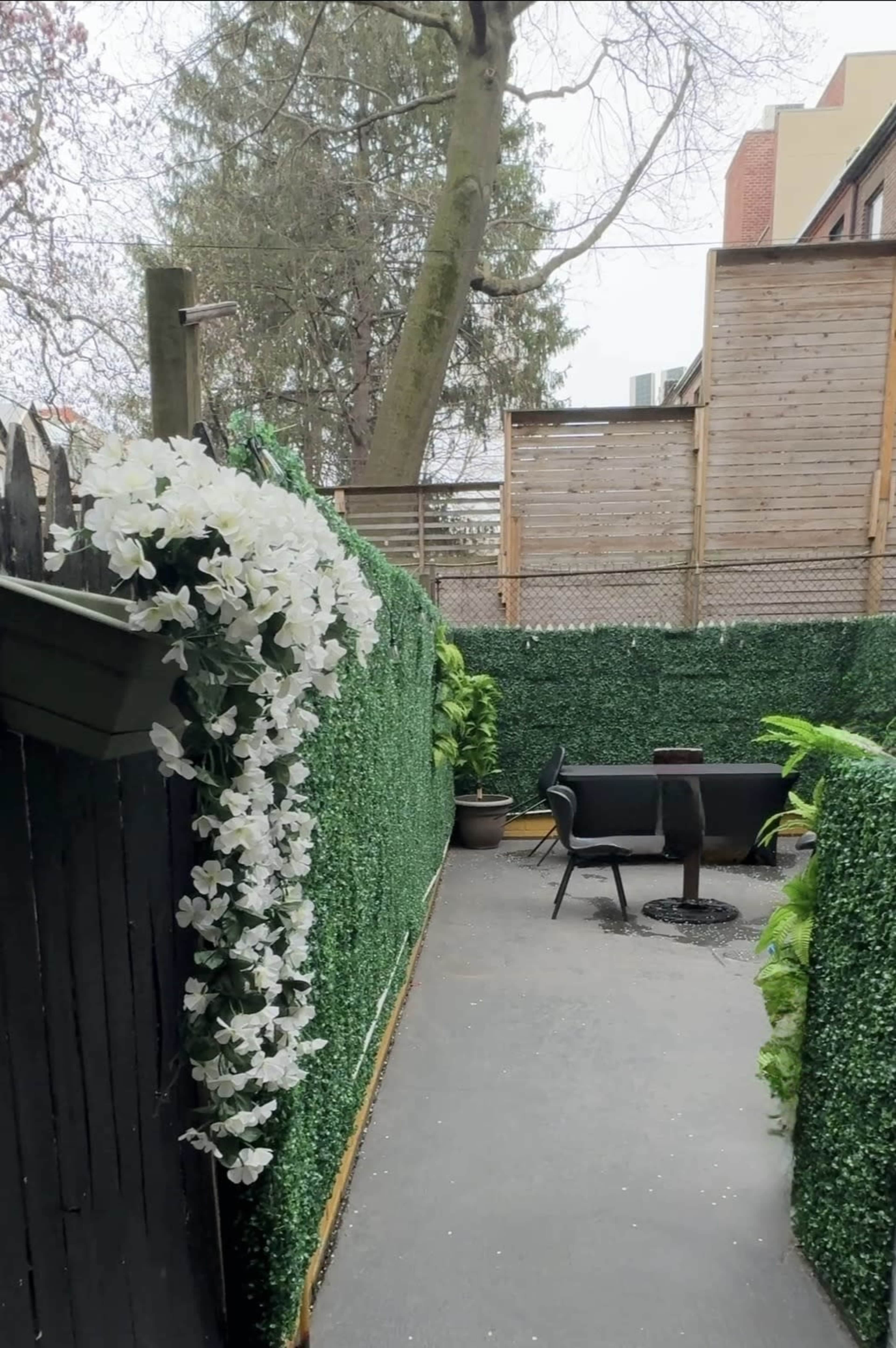 The image shows a narrow outdoor space enclosed by artificial greenery, featuring potted plants and patio furniture.