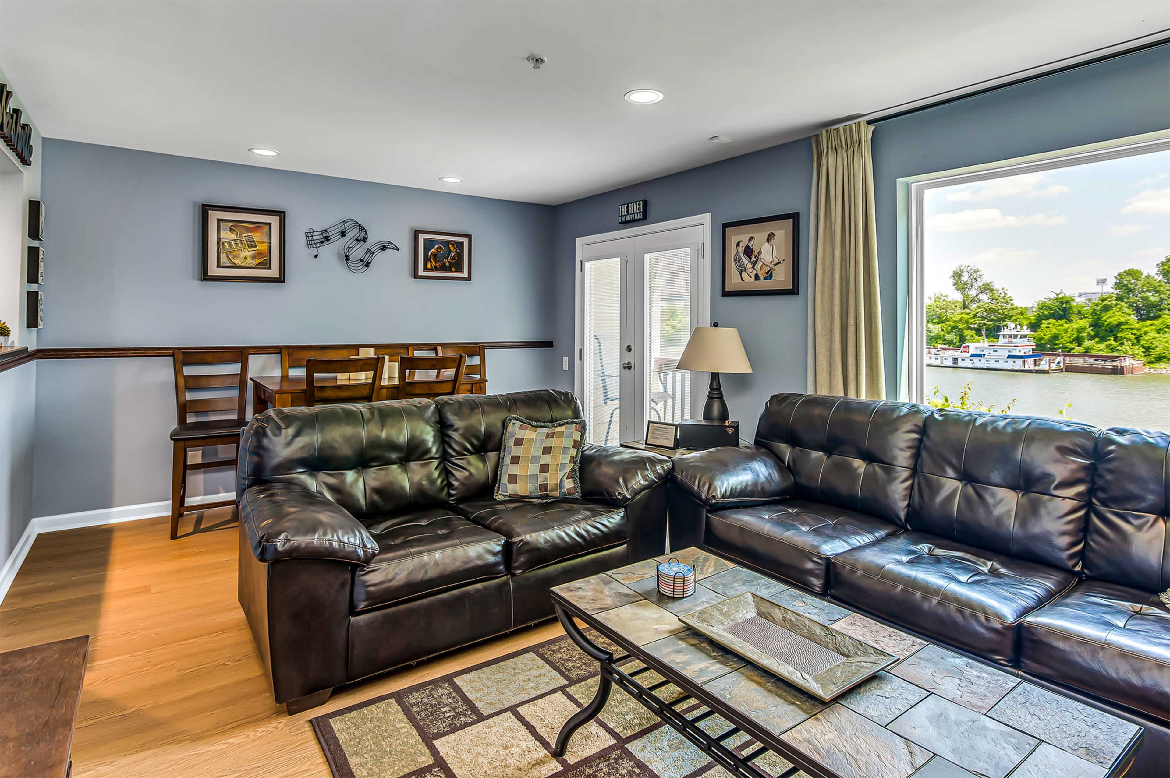 The image shows a living room with black leather sofas, a coffee table, and a view of a river through large windows.