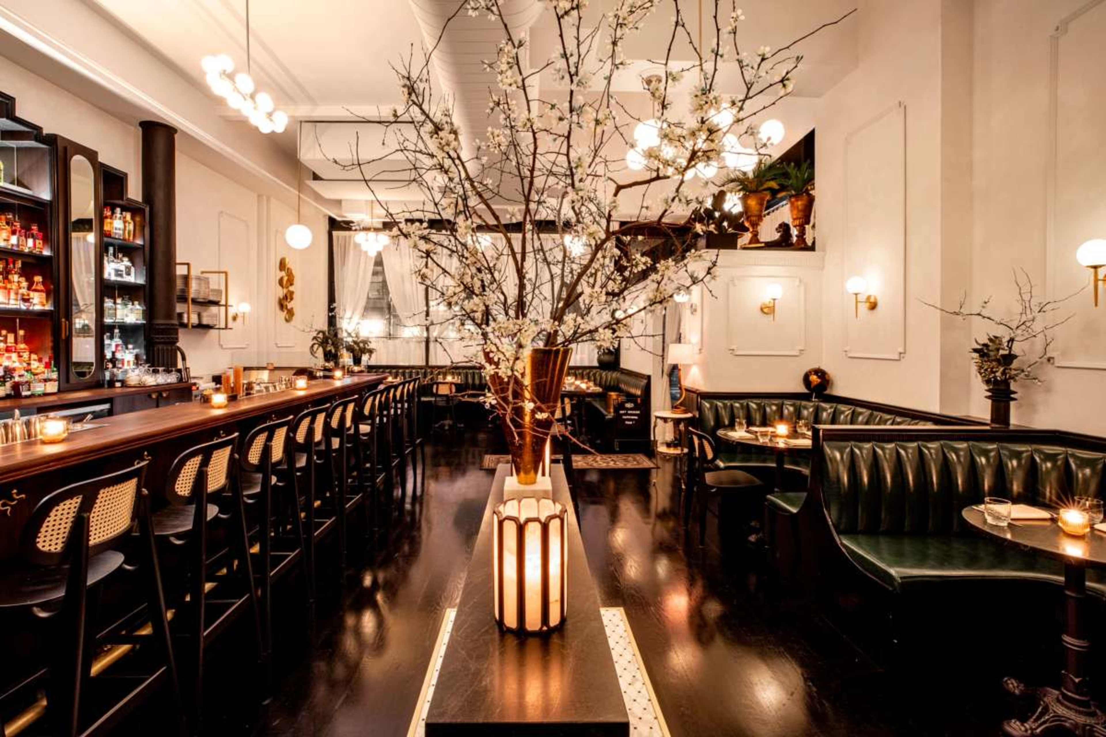 The image showcases a stylish restaurant with a central bar, green leather seating, and decorative branches arranged in a vase.