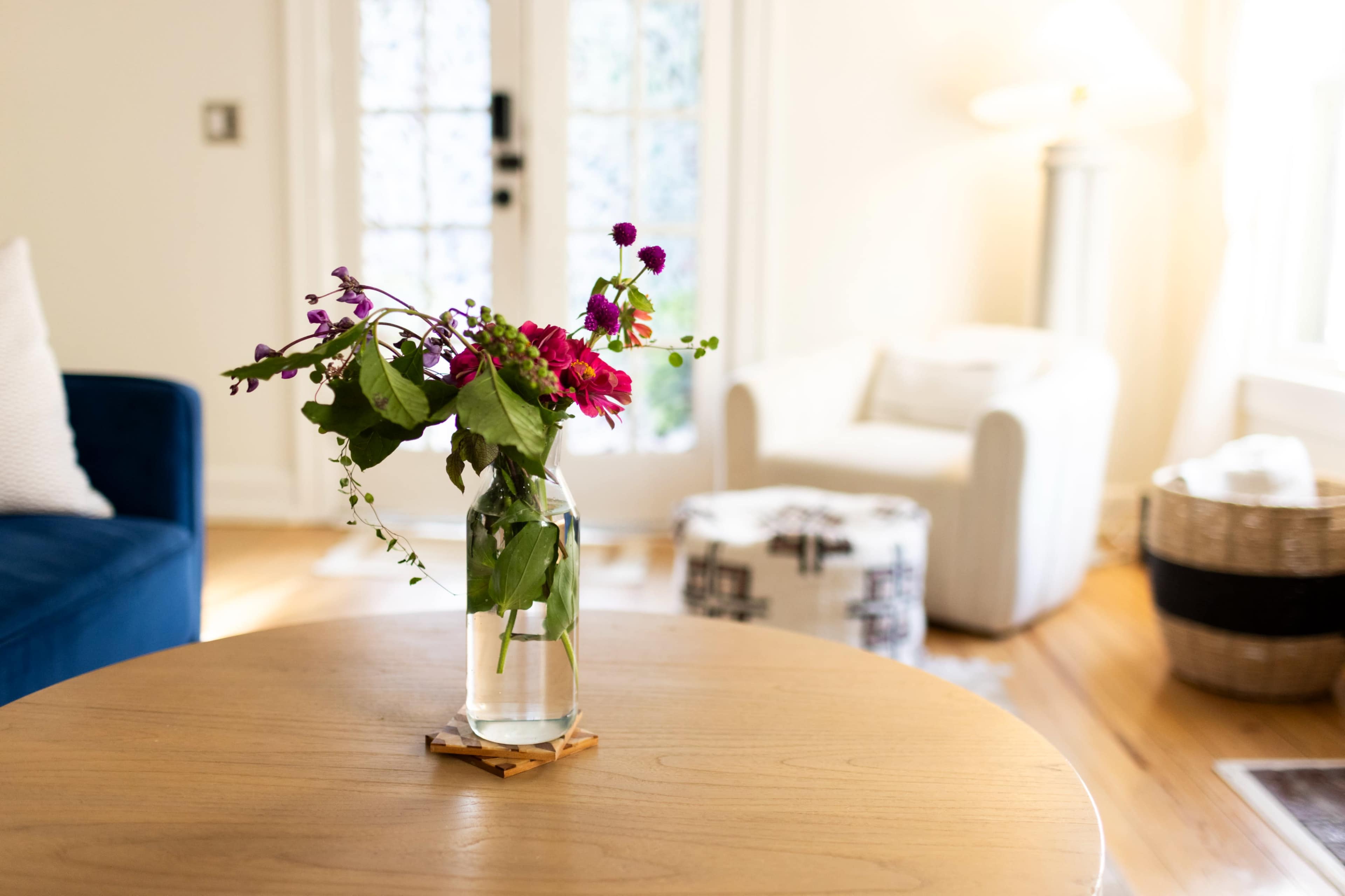 A vase of colorful flowers sits on a wooden table in a brightly lit living room with a blue couch and a white armchair in the background.