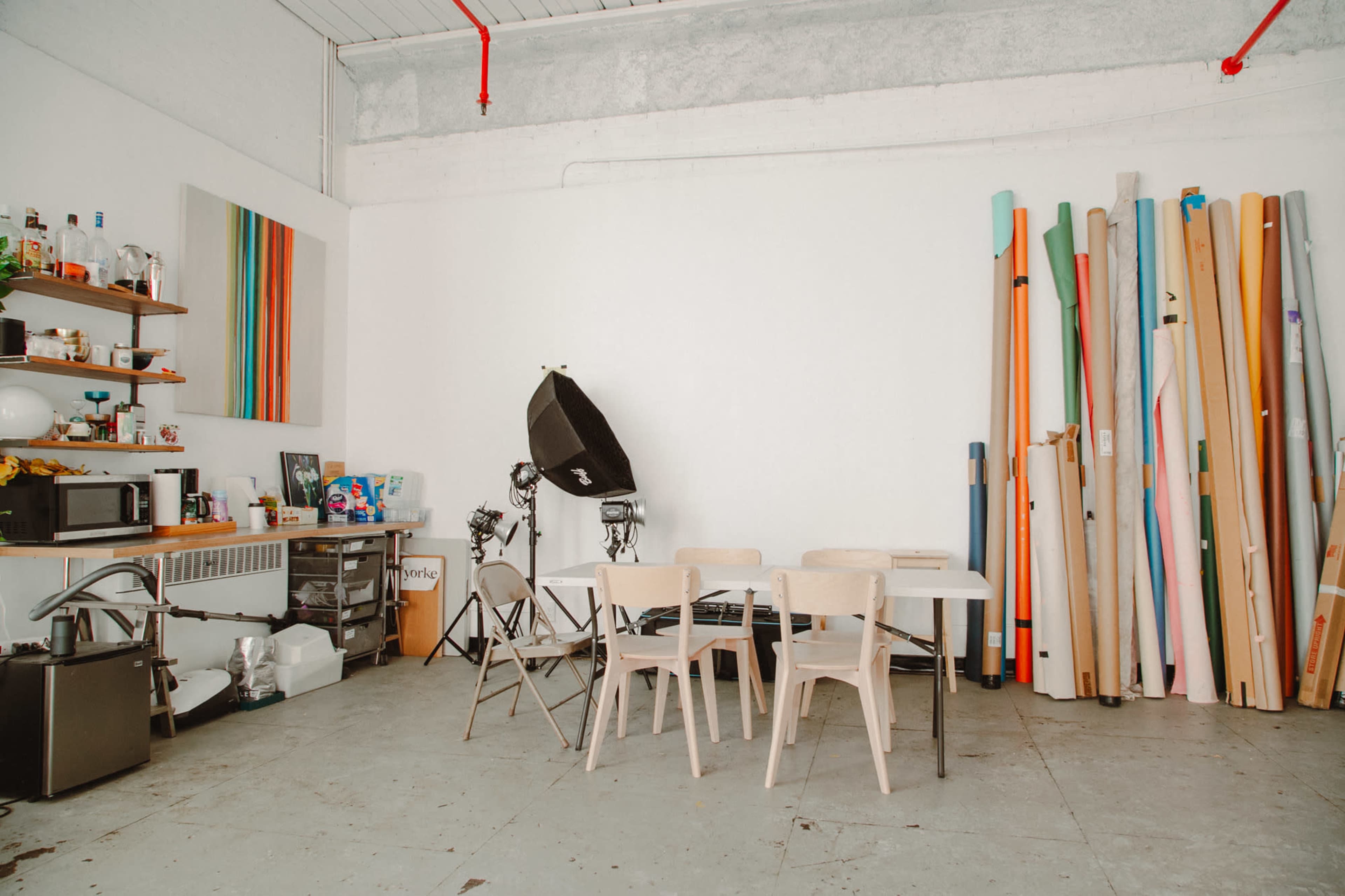 The image shows a spacious studio with a table and chairs, shelves filled with various items, and a collection of rolled materials leaning against the wall.