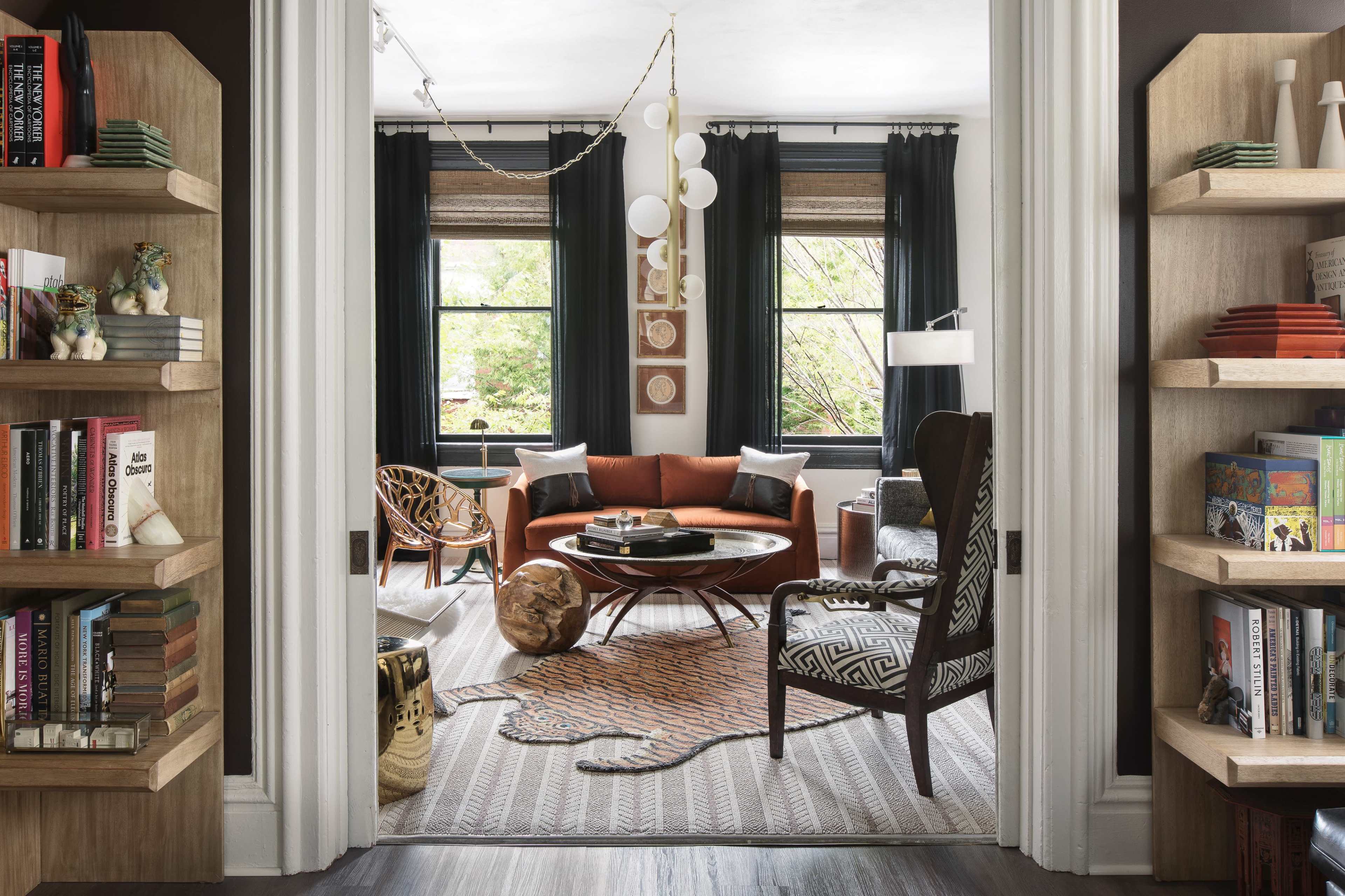 A cozy living room features a circular coffee table surrounded by an orange sofa, with bookshelves and large windows framing the space.