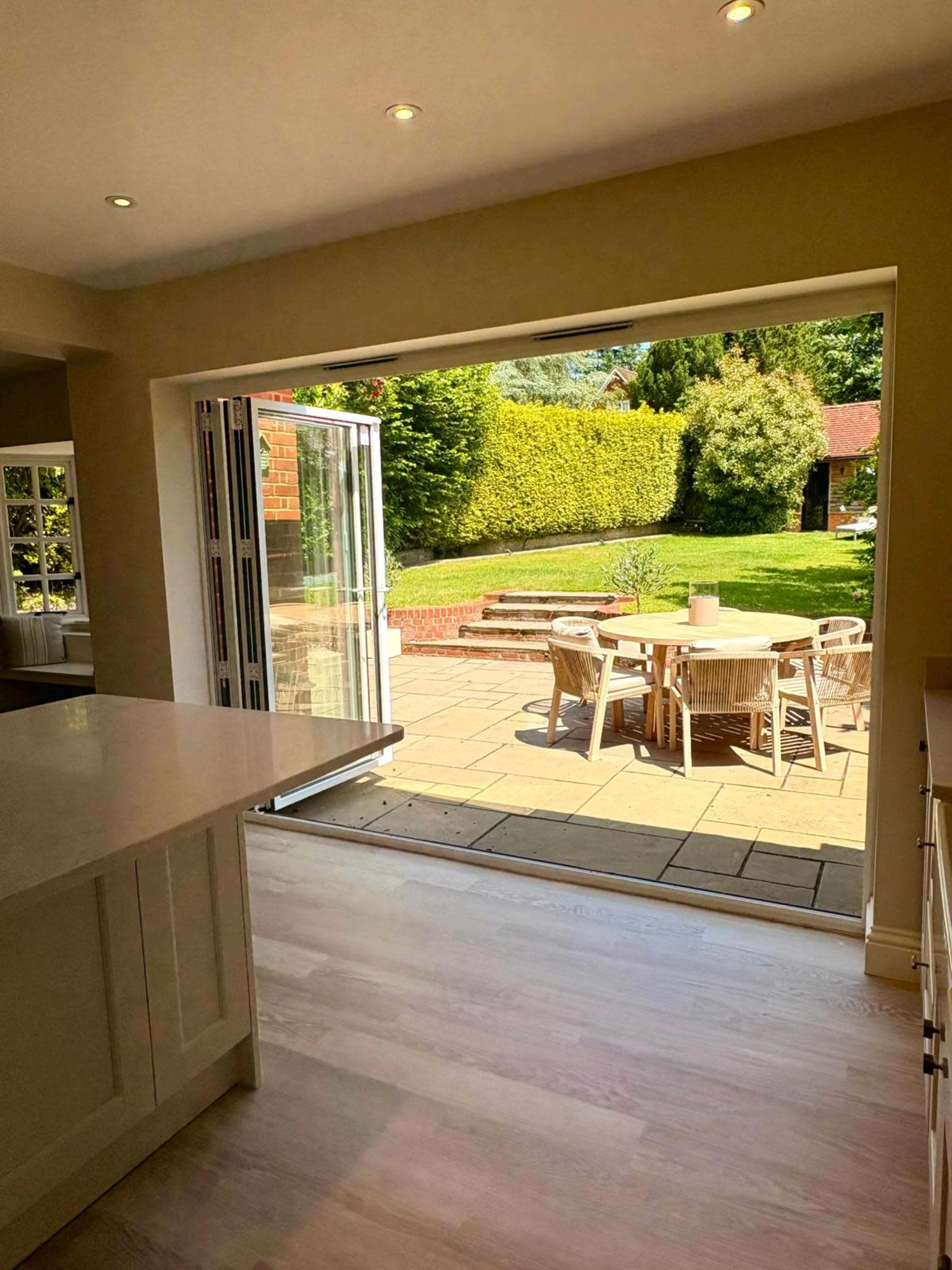 A modern kitchen opens up to a patio area with a dining table and lush greenery beyond.