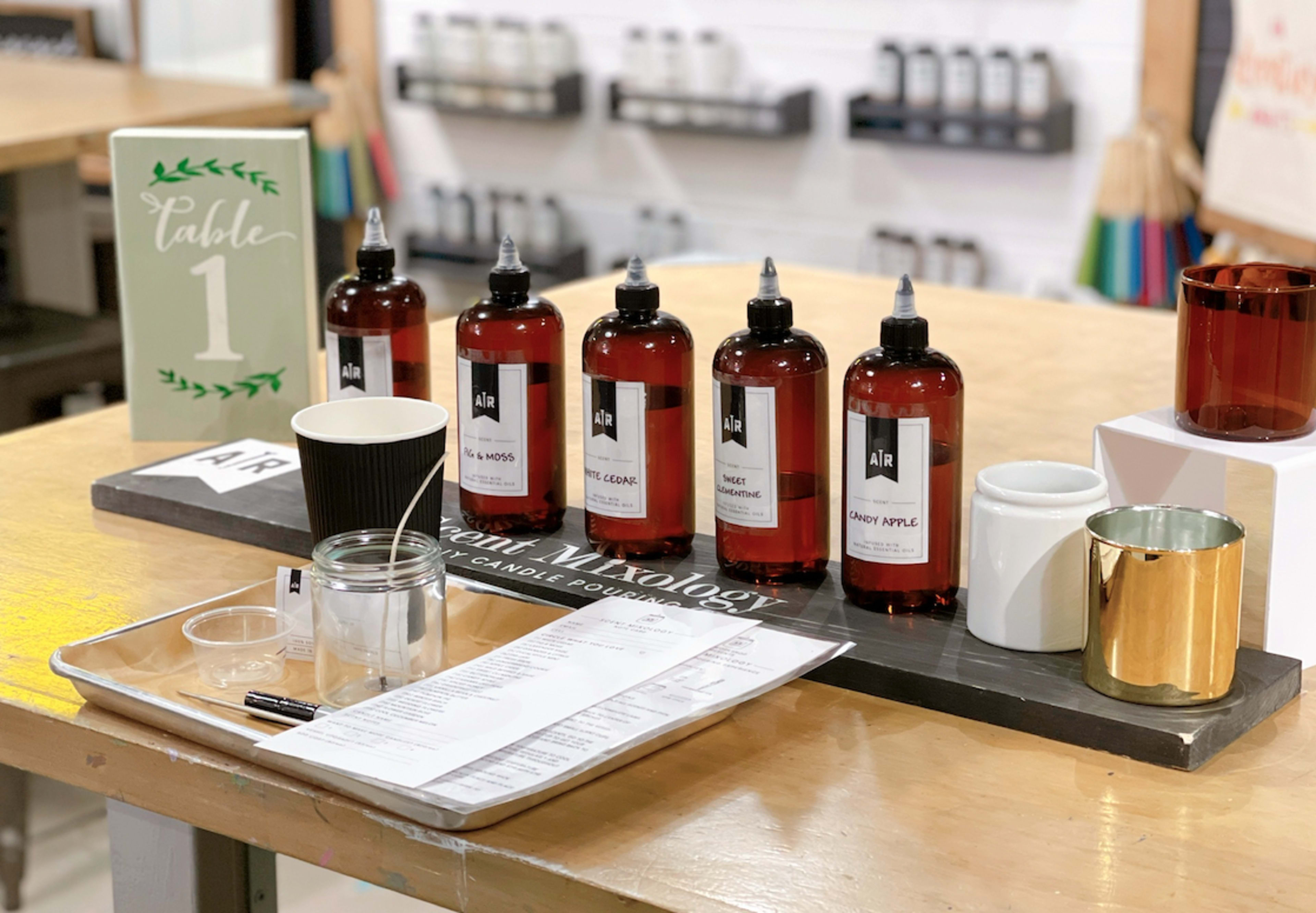 A setup for a candle-making station featuring several bottles of fragrance oils, a table number, a notepad, and various containers.