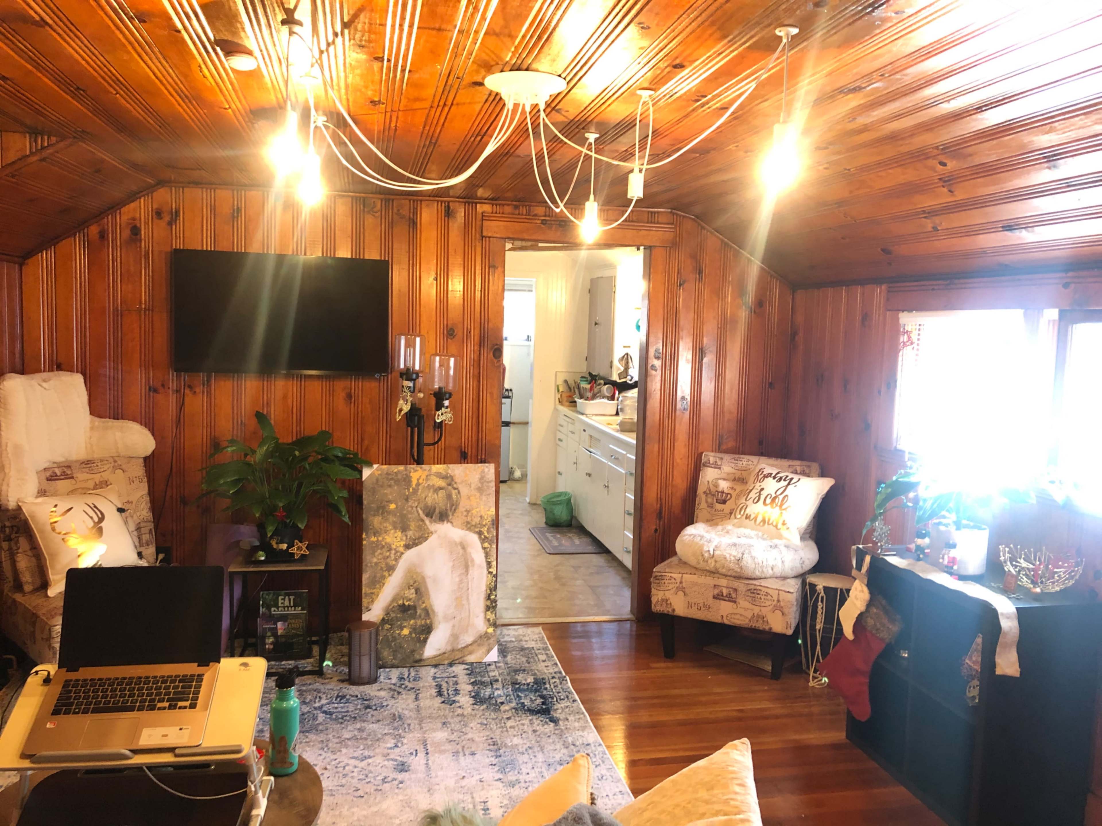 The room features wooden paneled walls, a small seating area with a laptop, a large decorative painting, and an entryway leading to a kitchen visible in the background.