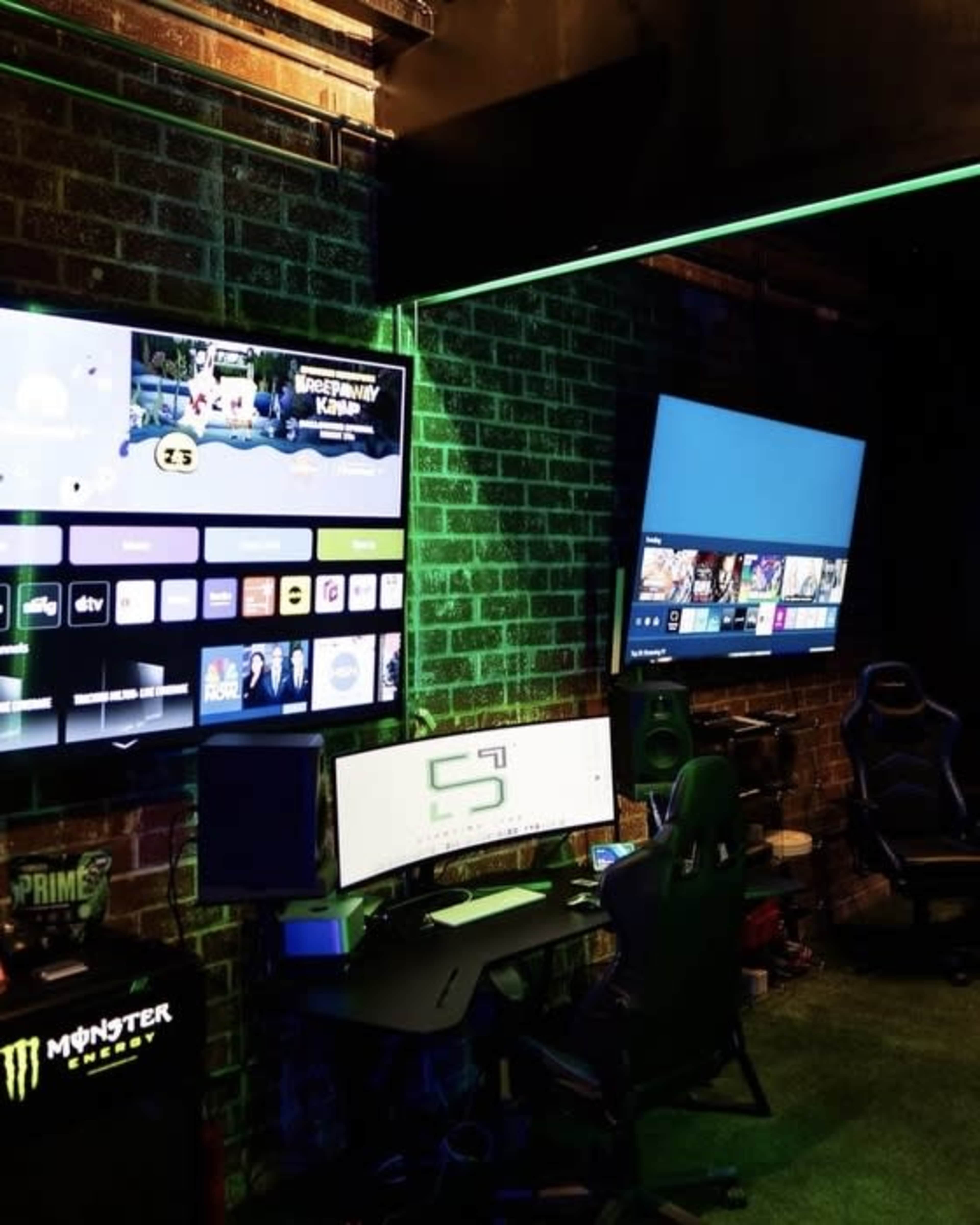 The image shows a gaming setup with two large screens mounted on a brick wall, featuring a desk and gaming chairs in front of them.
