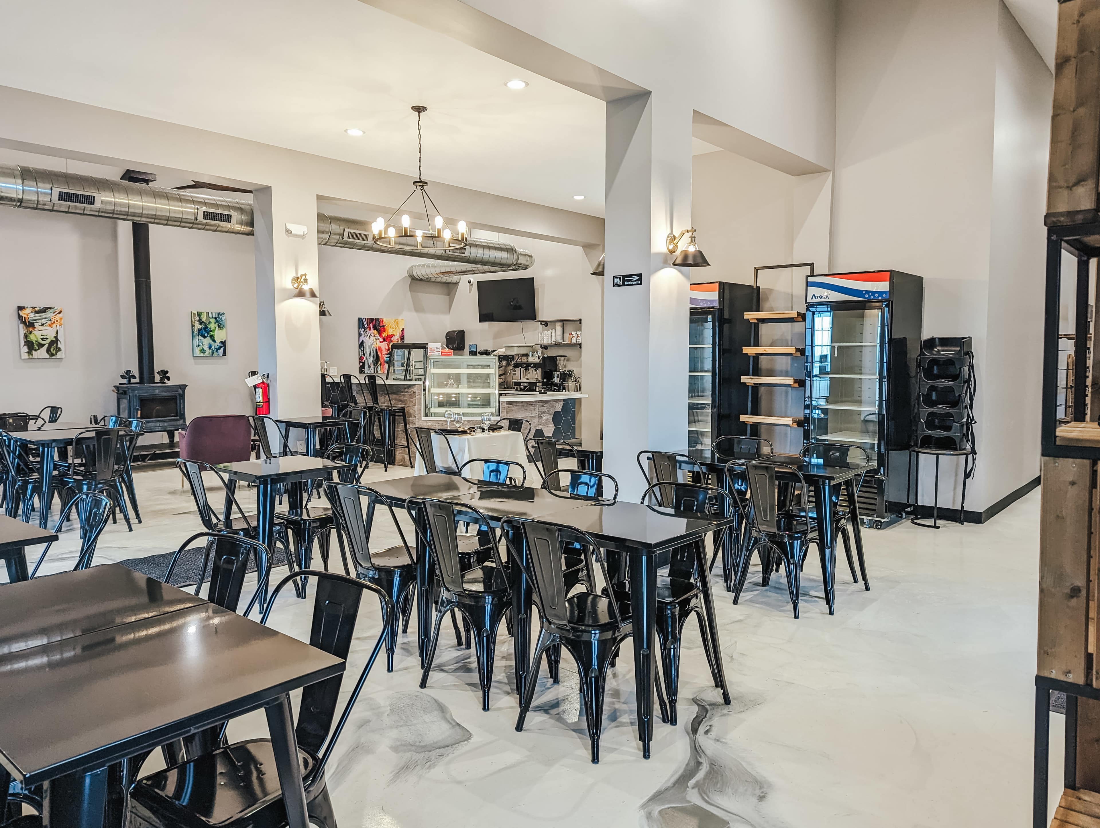 The image shows a modern café with black metal tables and chairs arranged in an open space featuring a display case and shelves in the background.