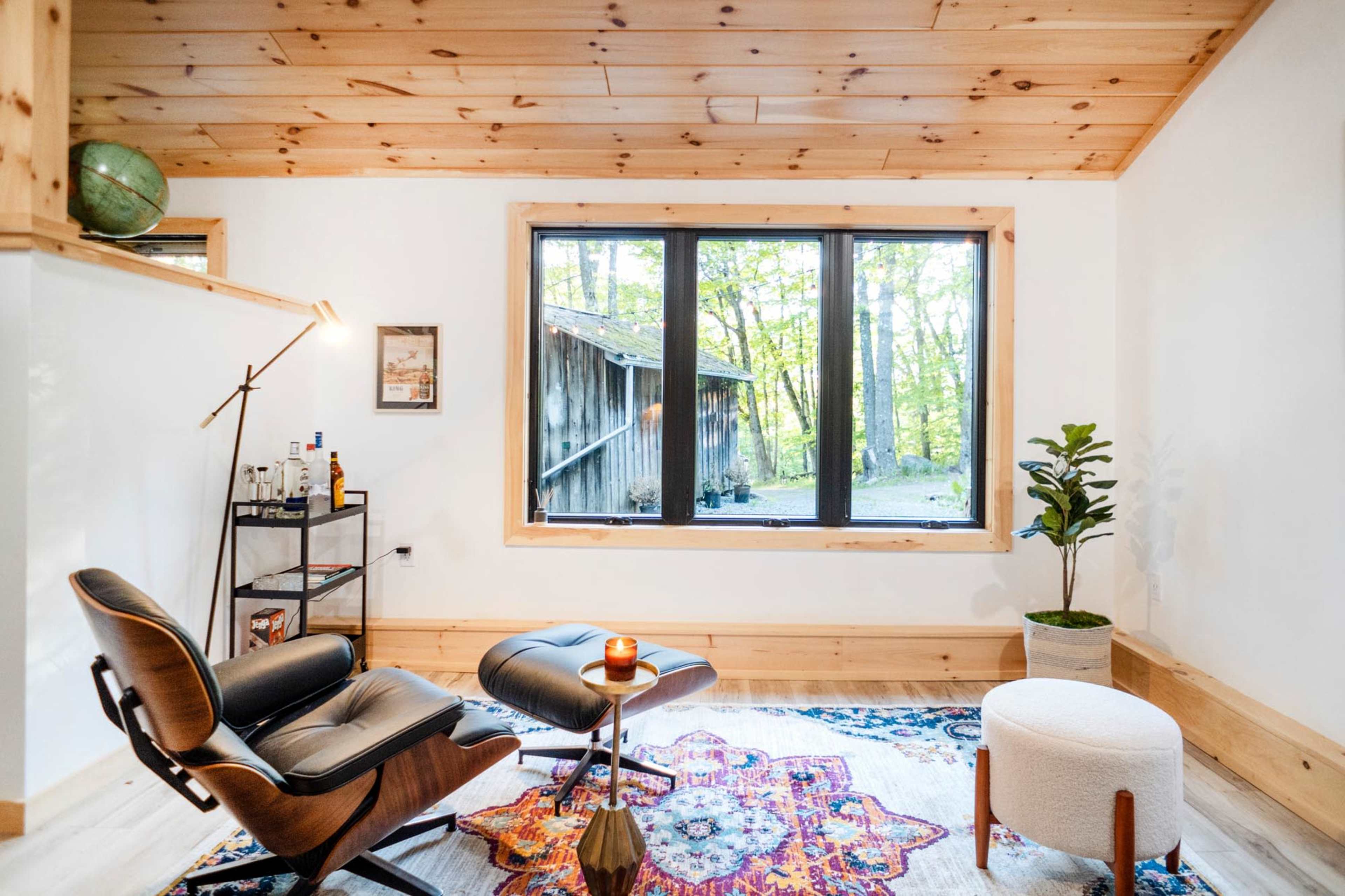 A living space features a modern chair and ottoman, a small side table, and a large window overlooking a wooded area.