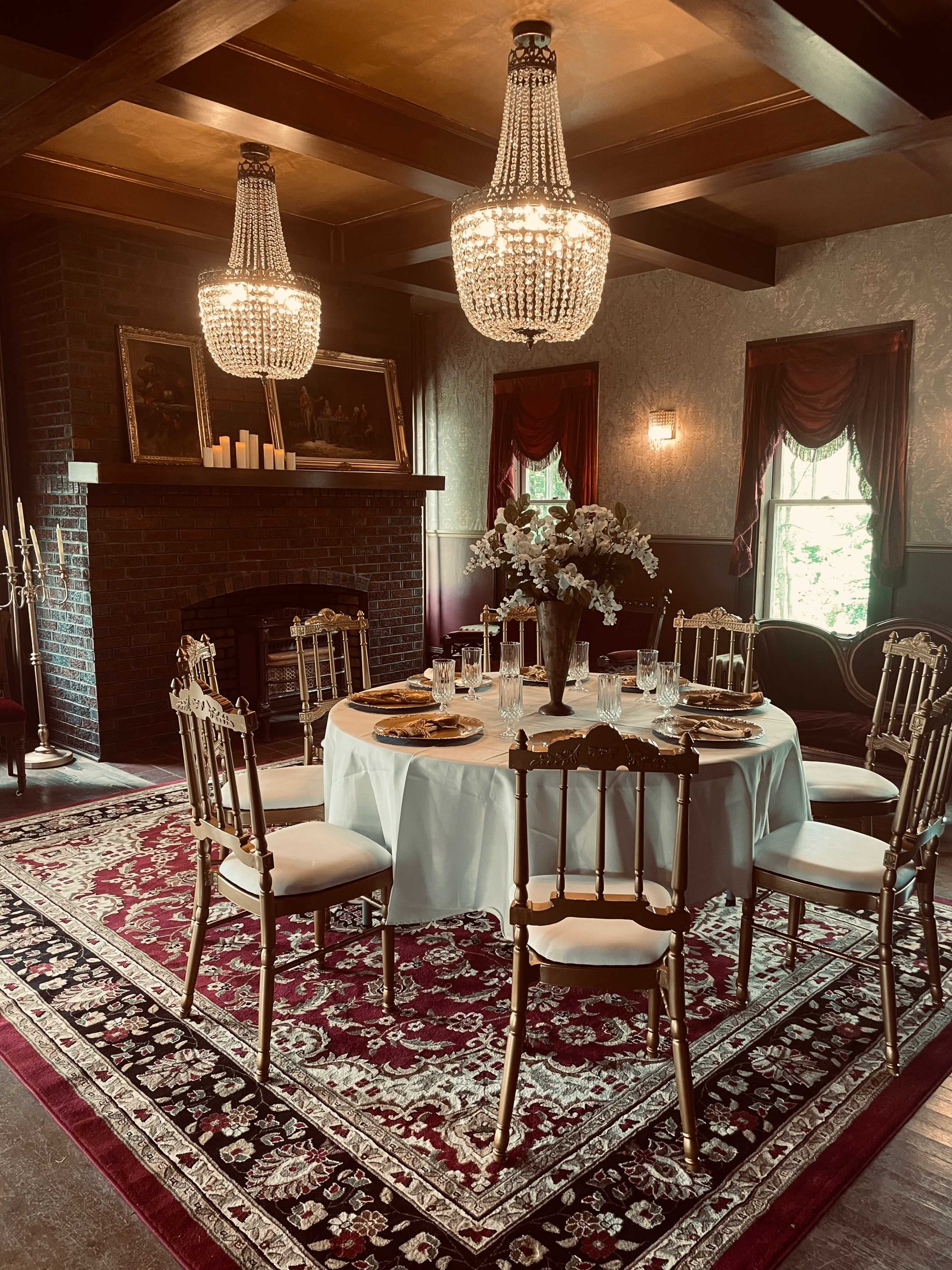 A round dining table set with plates and glasses is centered in an elegant dining room featuring chandeliers, a fireplace, and a decorative rug.