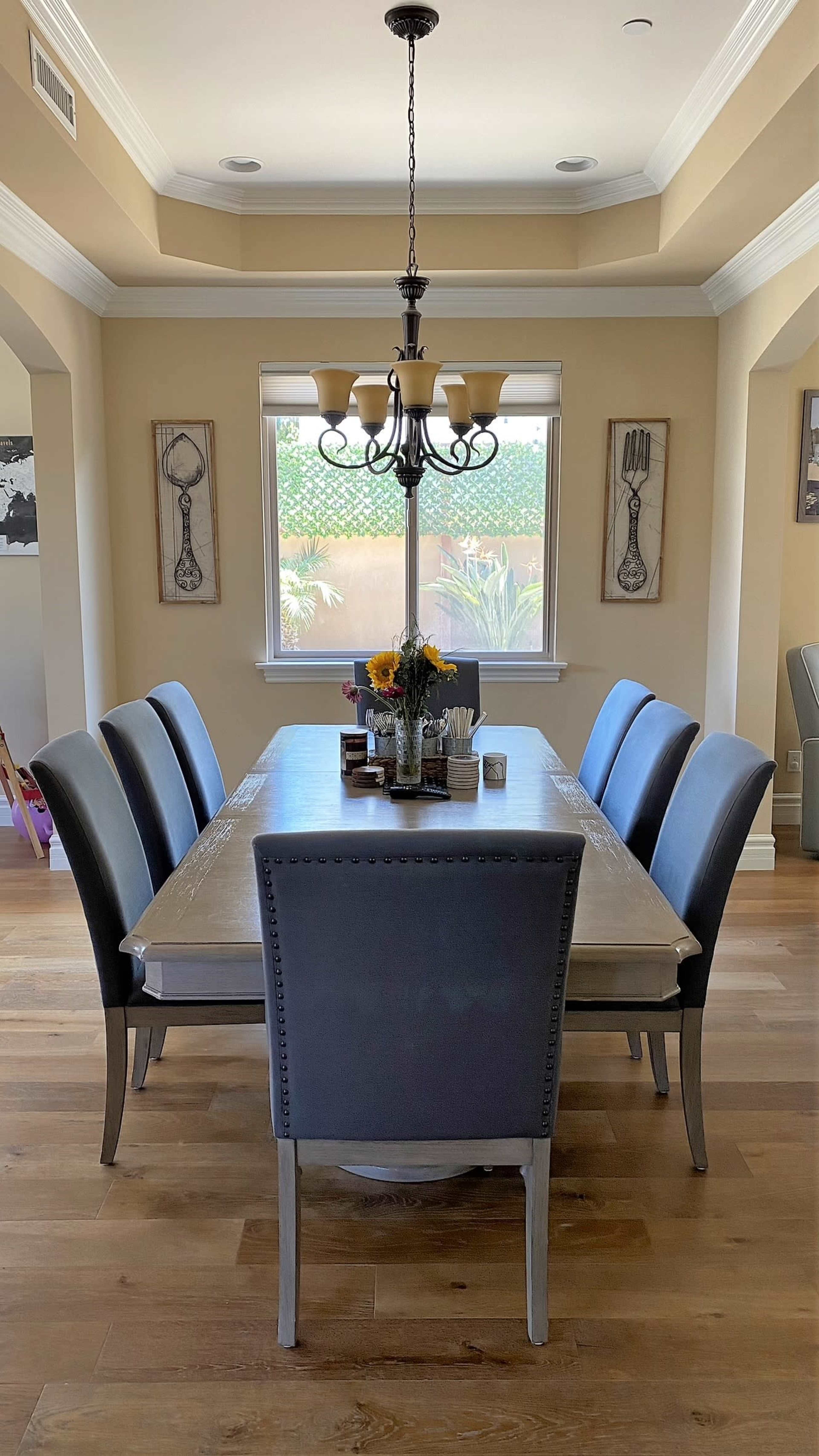 A dining room features a long table surrounded by six gray chairs, with a chandelier hanging above and a vase of sunflowers in the center.