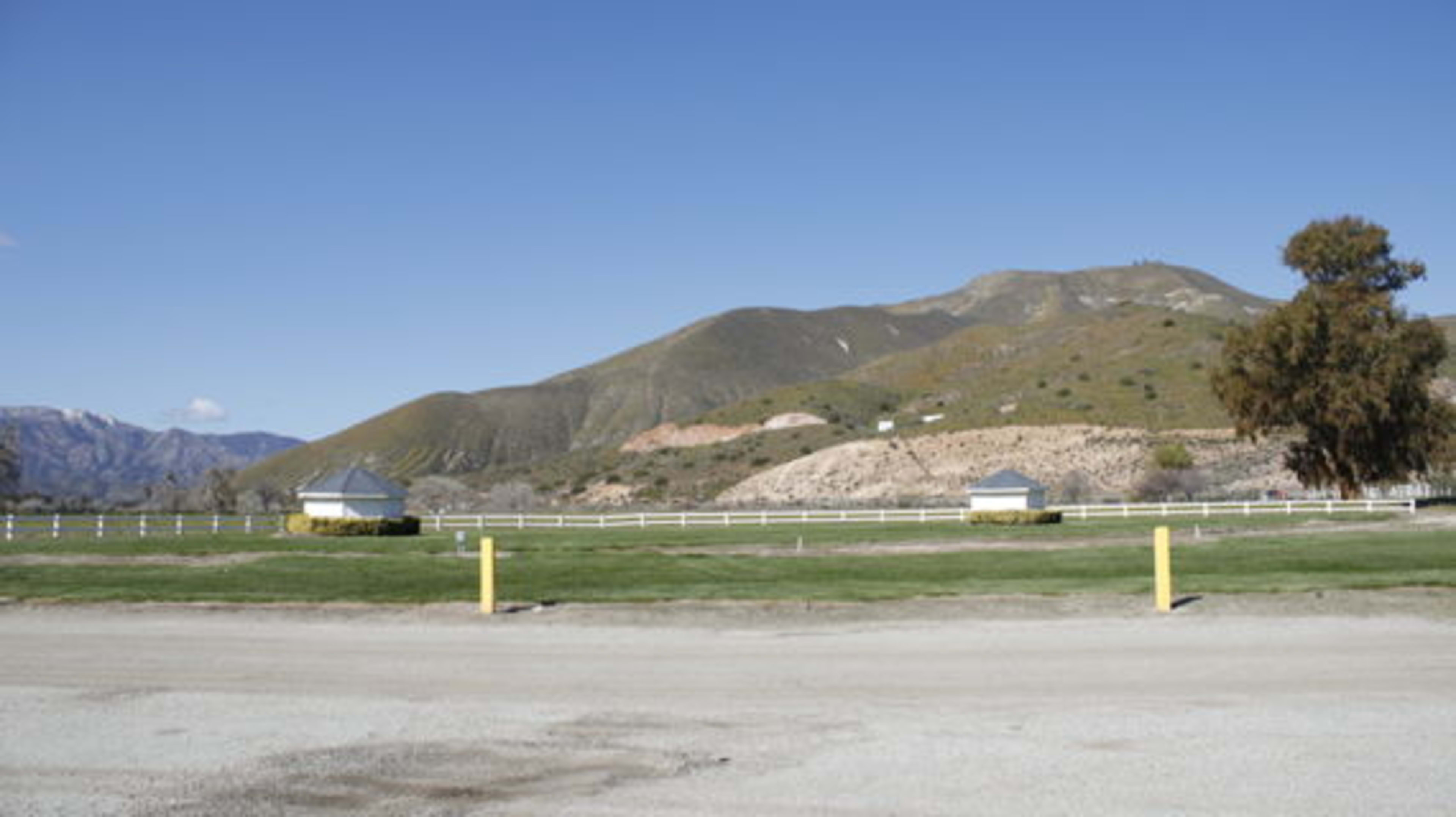 A grassy area with two small structures, surrounded by a white fence and a mountainous landscape in the background.