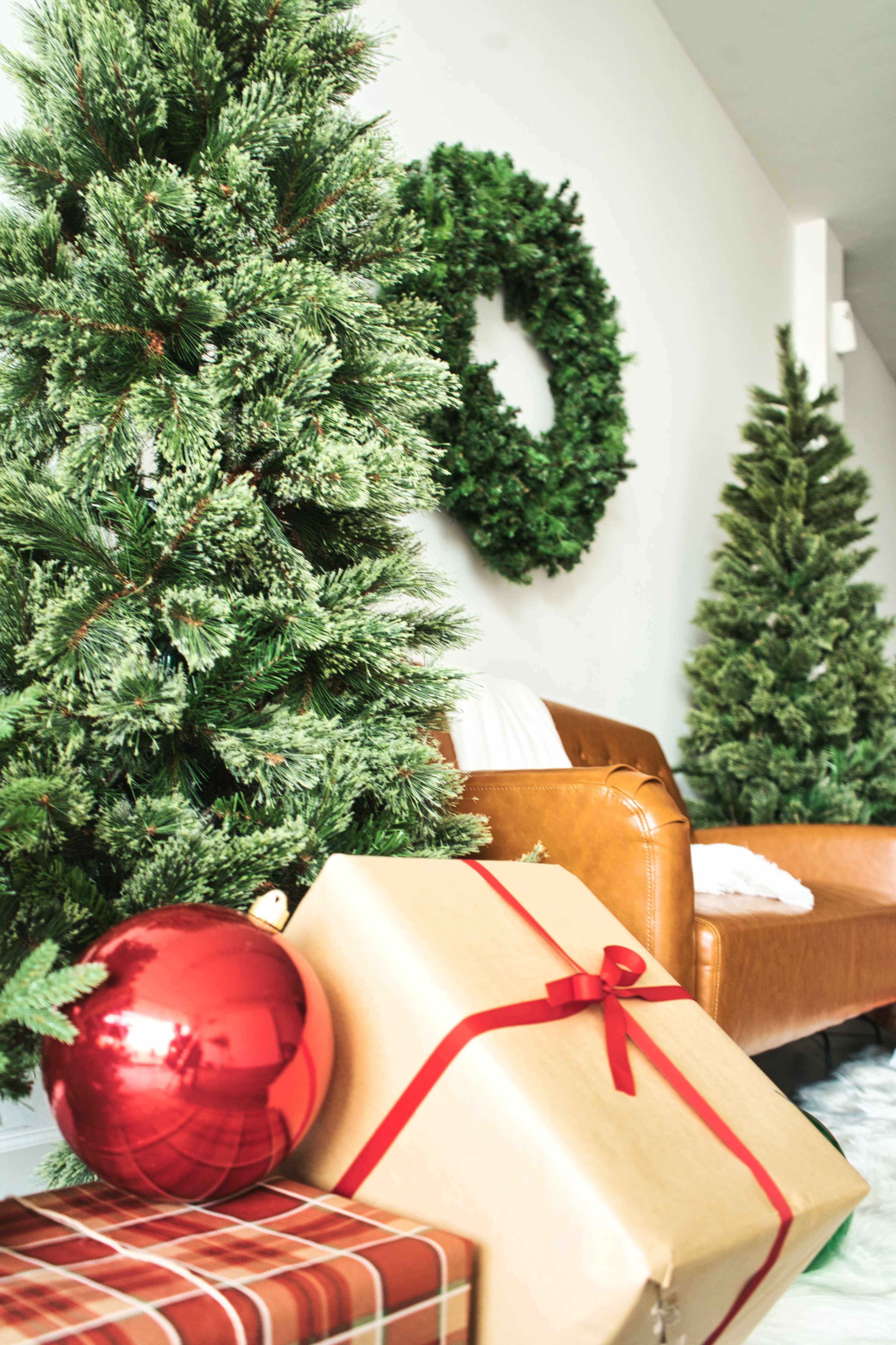 The image features a cozy living room decorated with two Christmas trees, a wreath on the wall, and stacked gifts beside a brown leather chair.