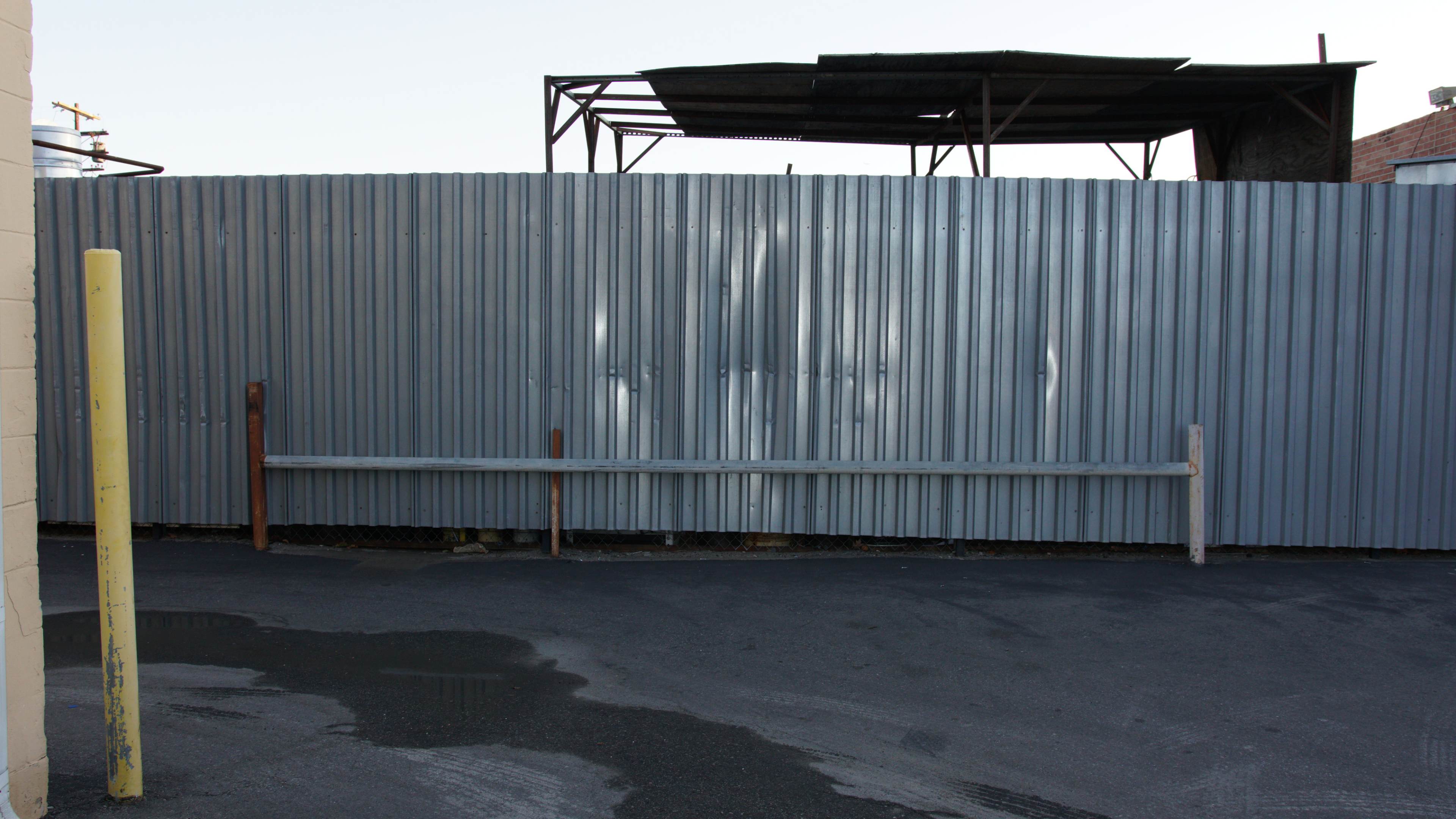A corrugated metal fence with a roof structure visible above it, set in an alleyway with wet pavement.