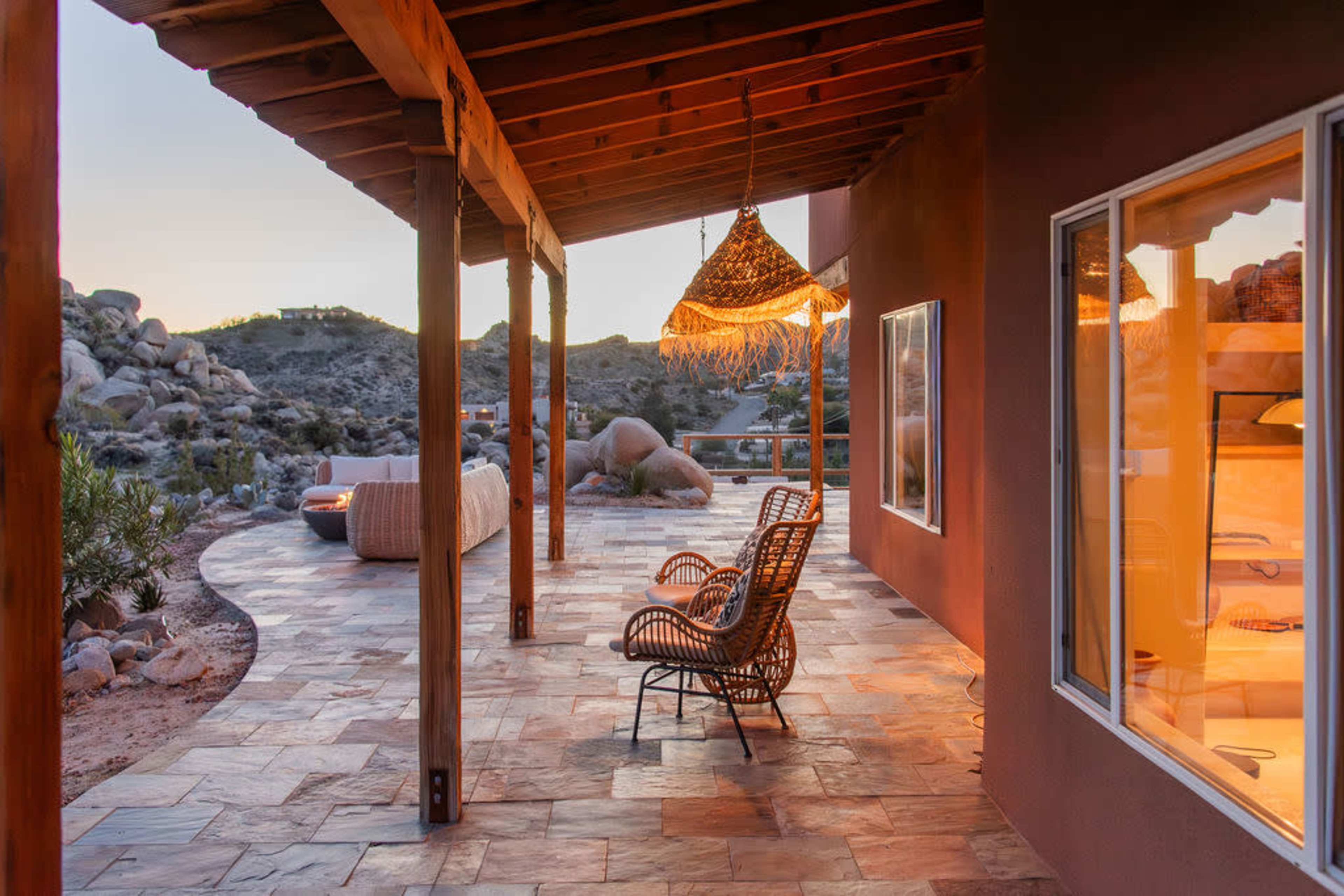 The image shows a patio area with a woven chair and a hanging light fixture, featuring a stone pathway and rugged hills in the background during sunset.