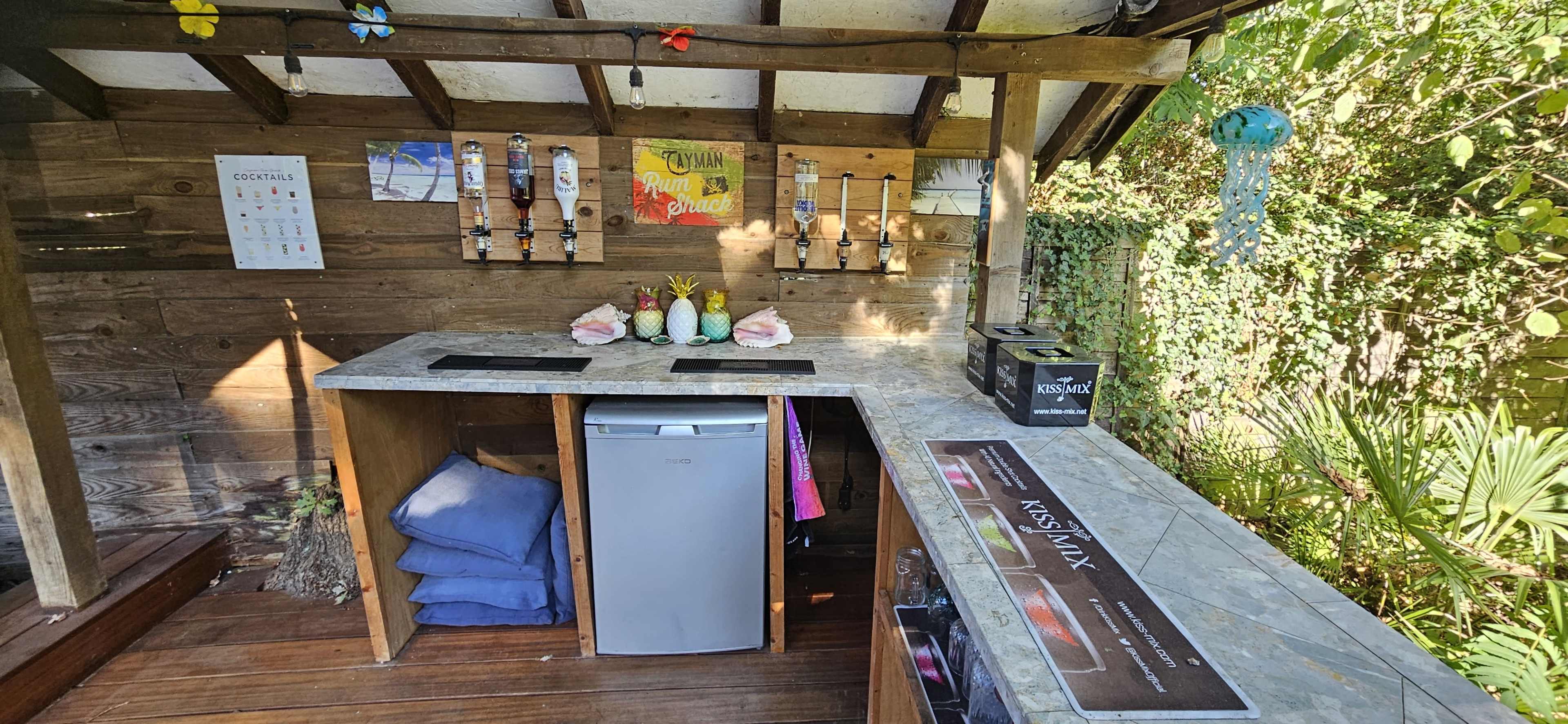 The image shows a wooden outdoor bar area with a stone countertop, featuring a small refrigerator and multiple drink dispensers mounted on the wall.