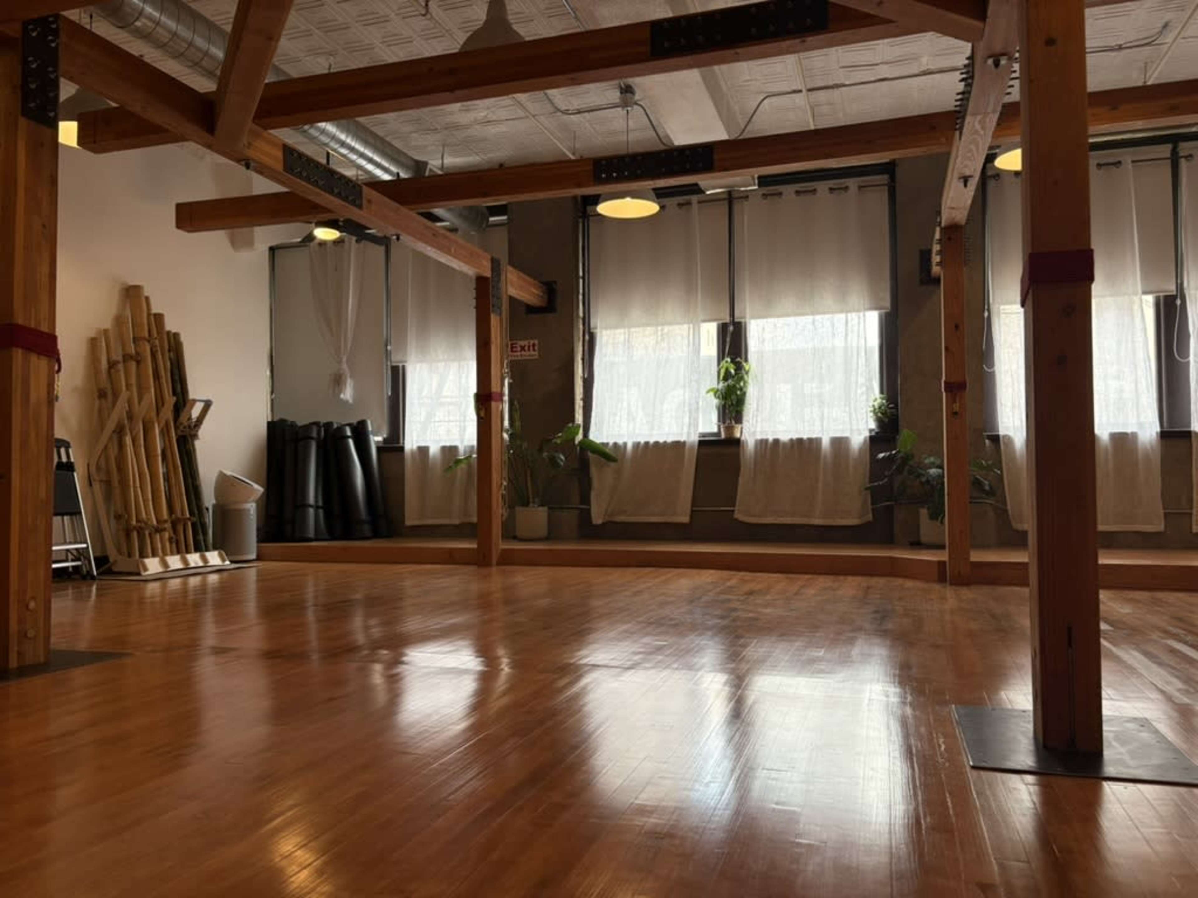 The image shows a spacious, well-lit wooden studio with large windows, wooden beams, and neatly arranged exercise equipment in the background.