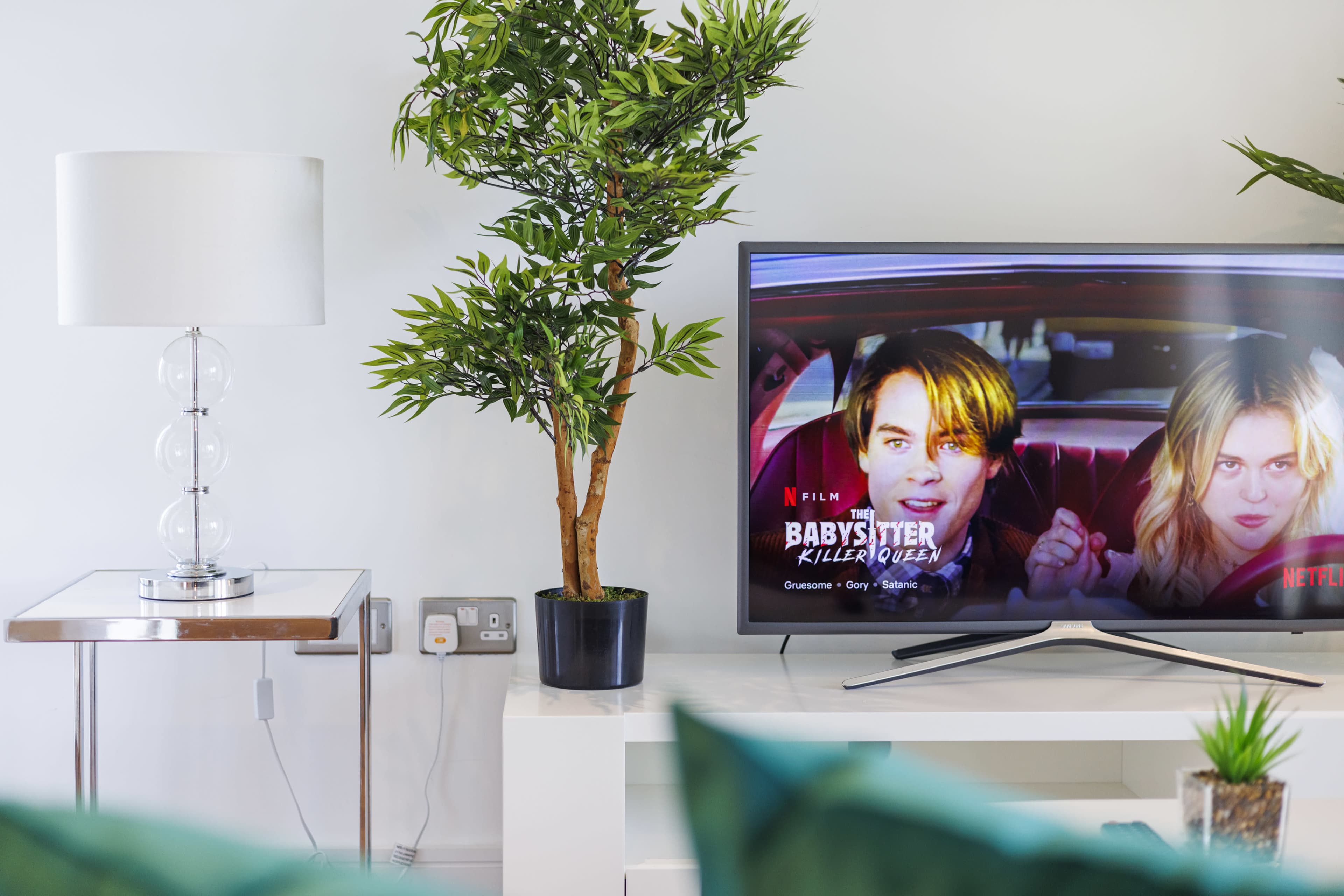 A television displaying a movie selection sits next to a potted plant and a lamp on a modern side table in a bright room.