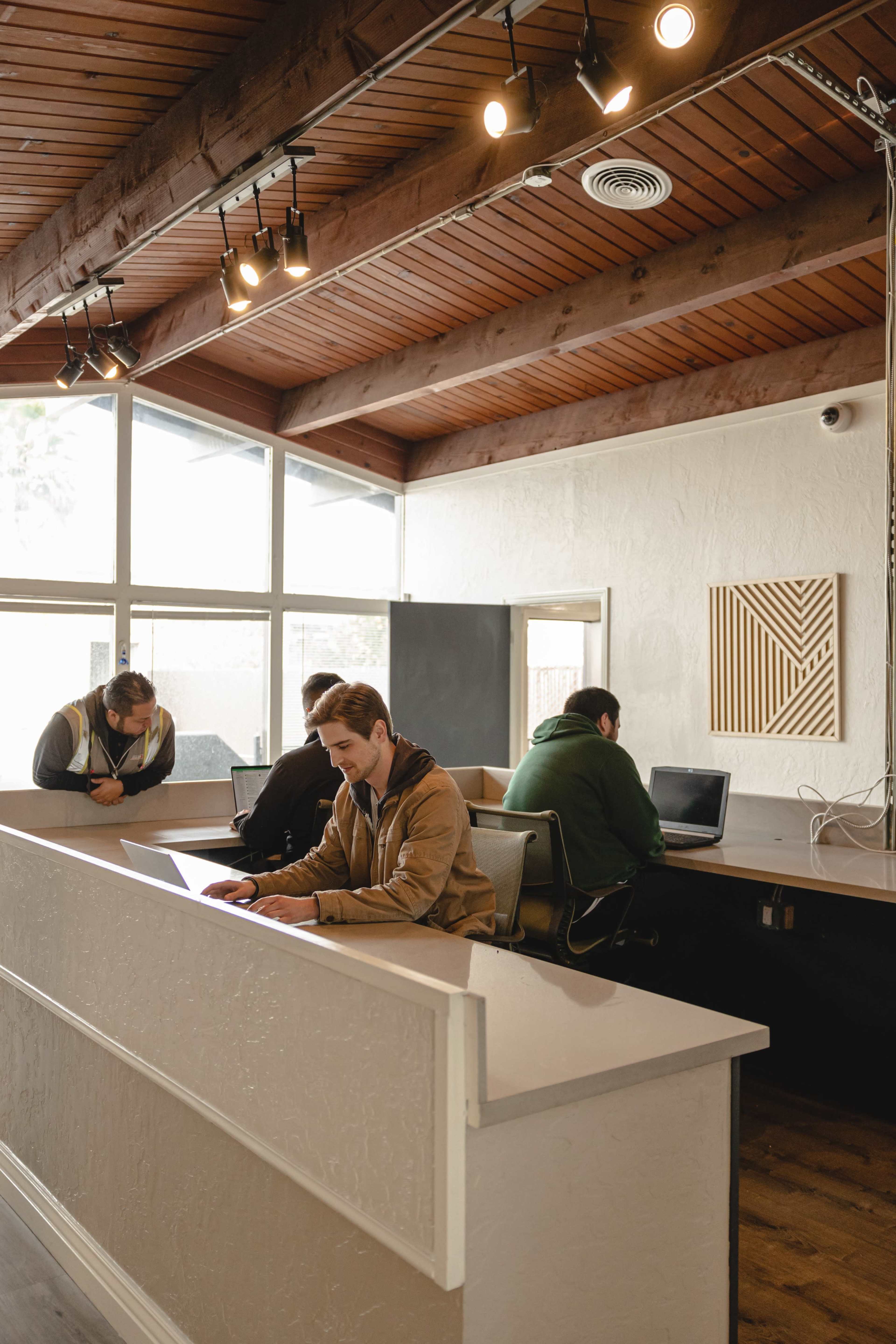 Four individuals are seated at a communal workspace, working on laptops in a well-lit room with wooden beams and large windows.