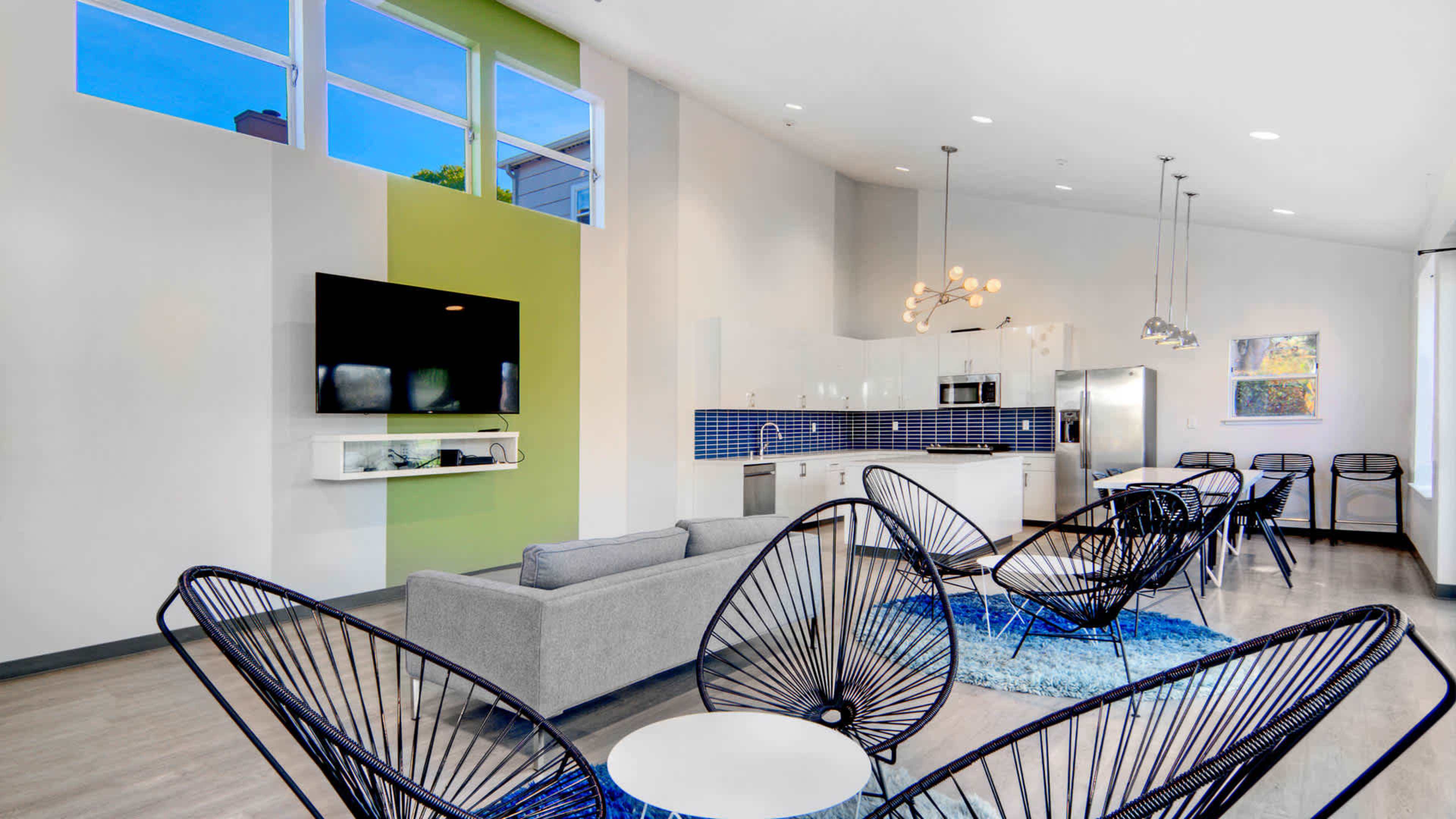 A bright and modern lounge area features a gray sofa, black chairs, a blue rug, and a kitchen with a blue backsplash and stainless-steel appliances.
