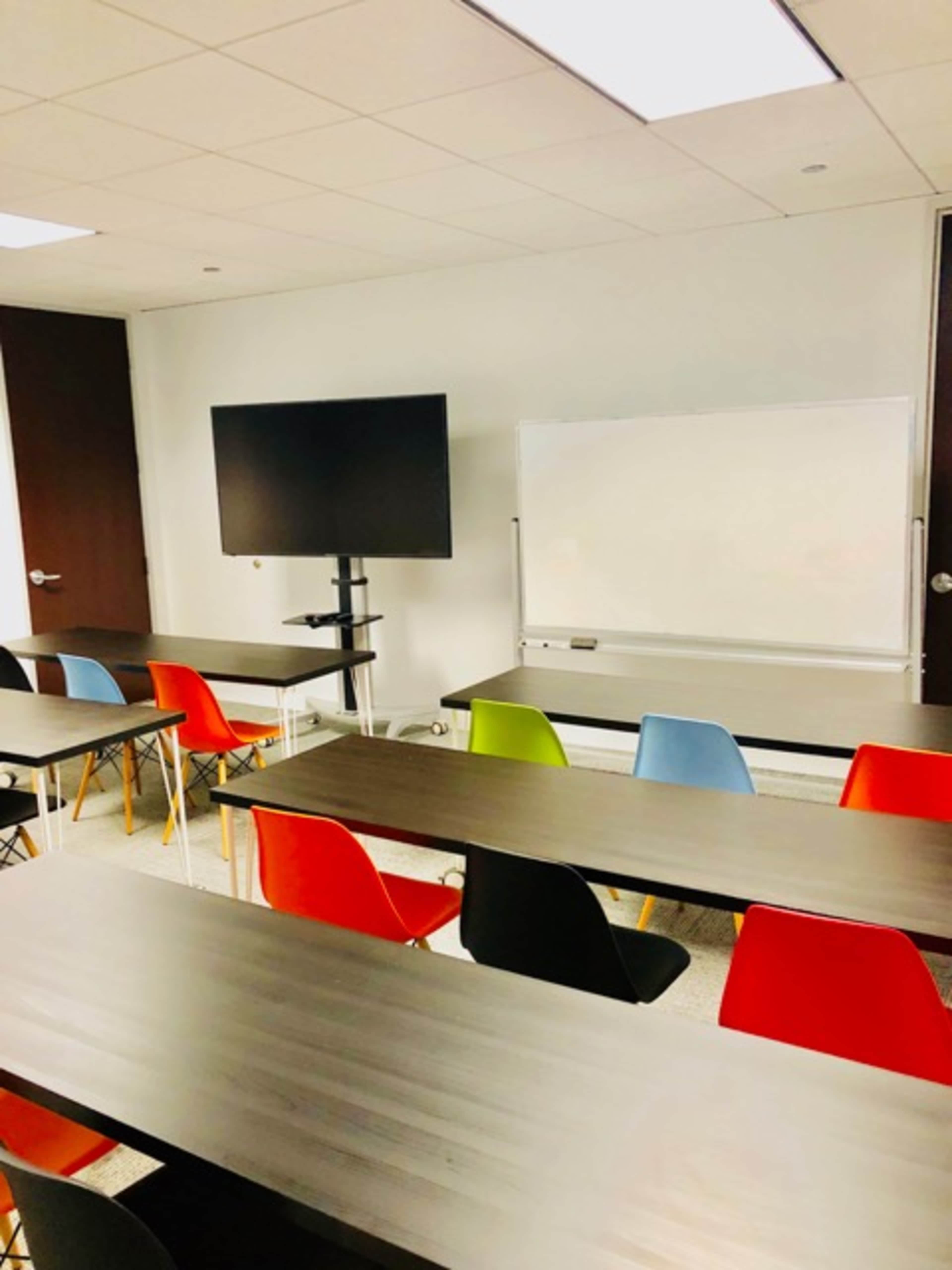 A classroom with multiple tables arranged in rows, colorful chairs, a whiteboard, and a television mounted on a stand.