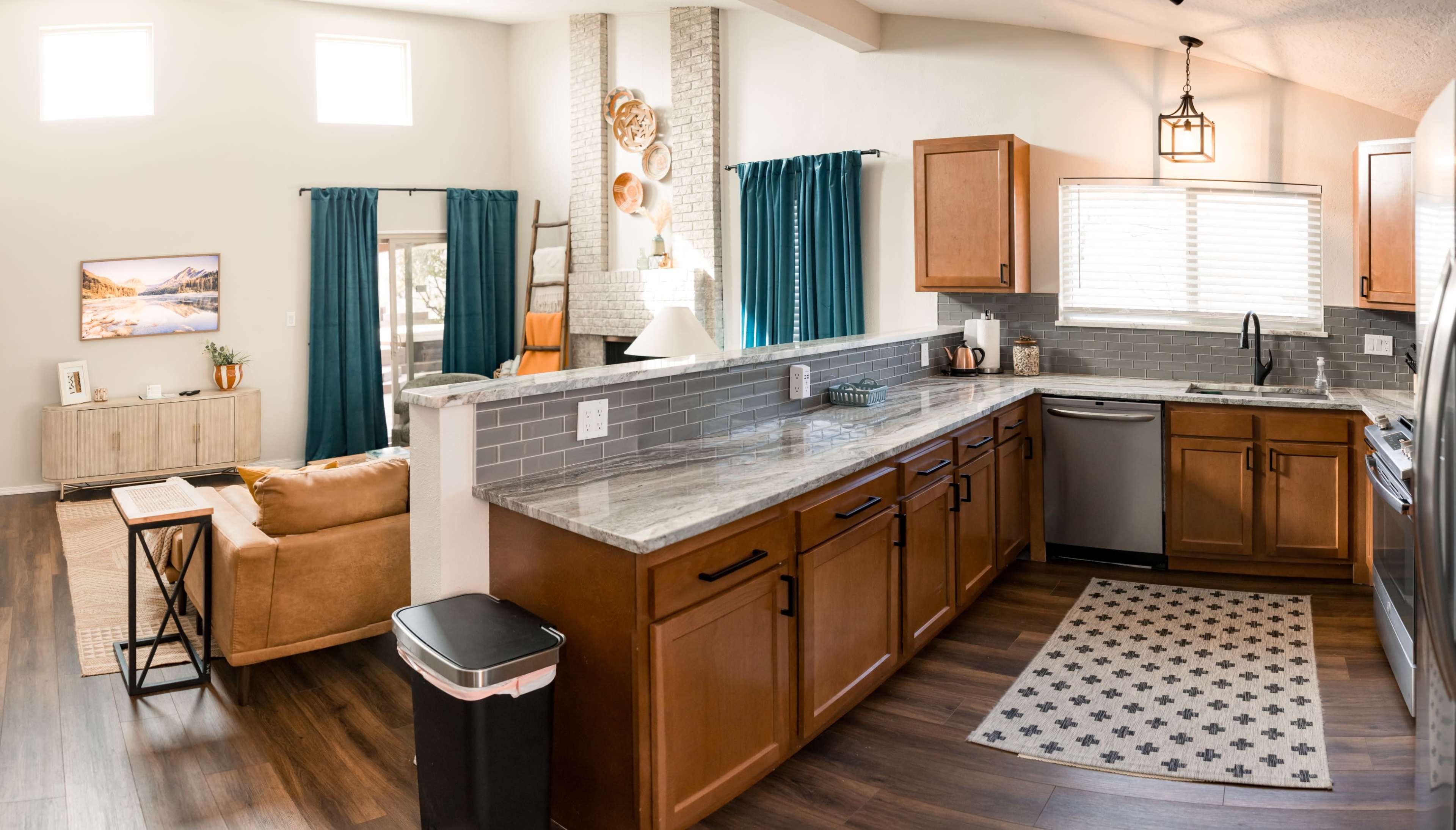 A modern kitchen with wooden cabinets, a stainless steel dishwasher, and a view of an open living area featuring a couch and a decorative fireplace.
