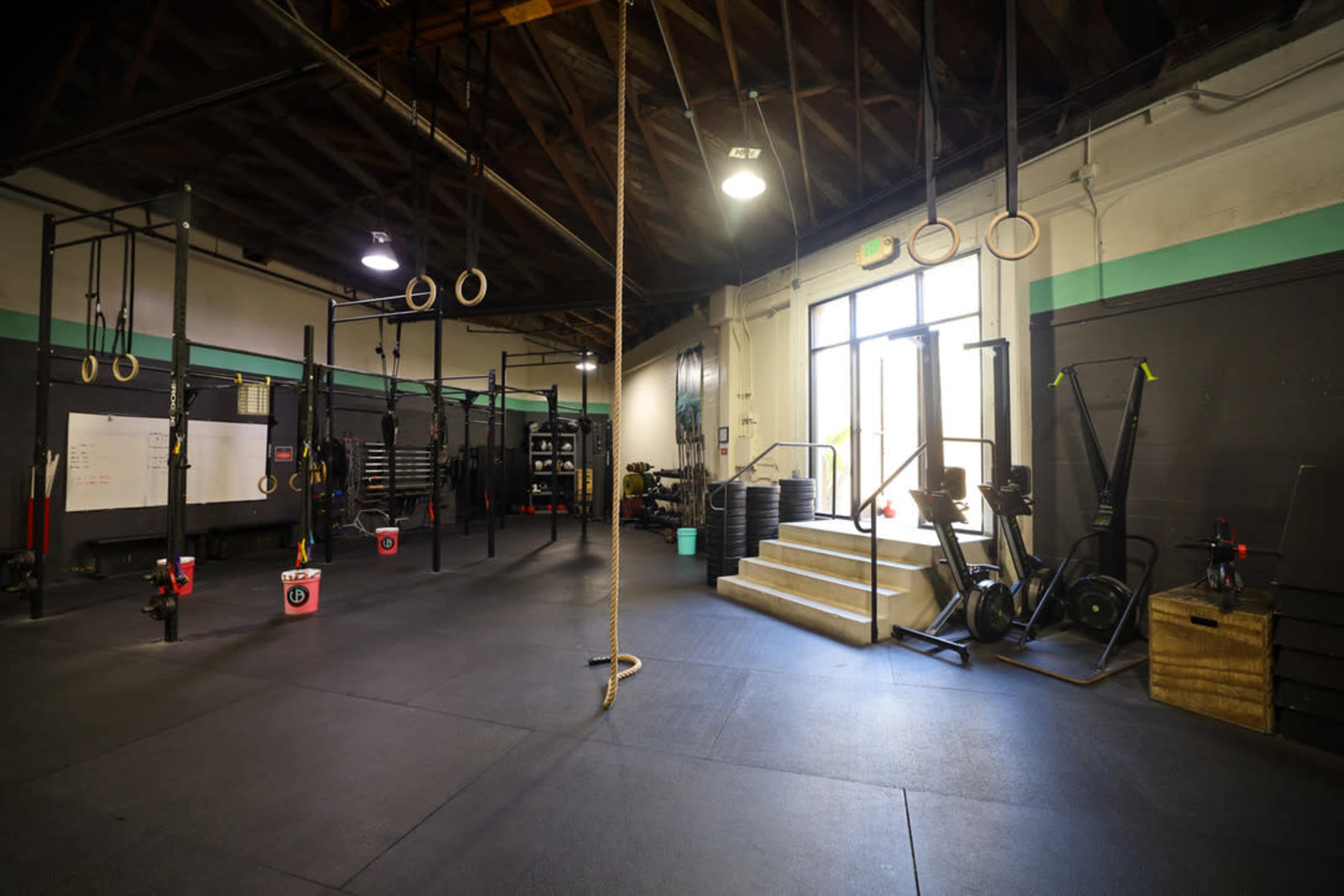 The image shows an empty gym with exercise equipment, including rings and rowing machines, and a set of stairs leading outside.