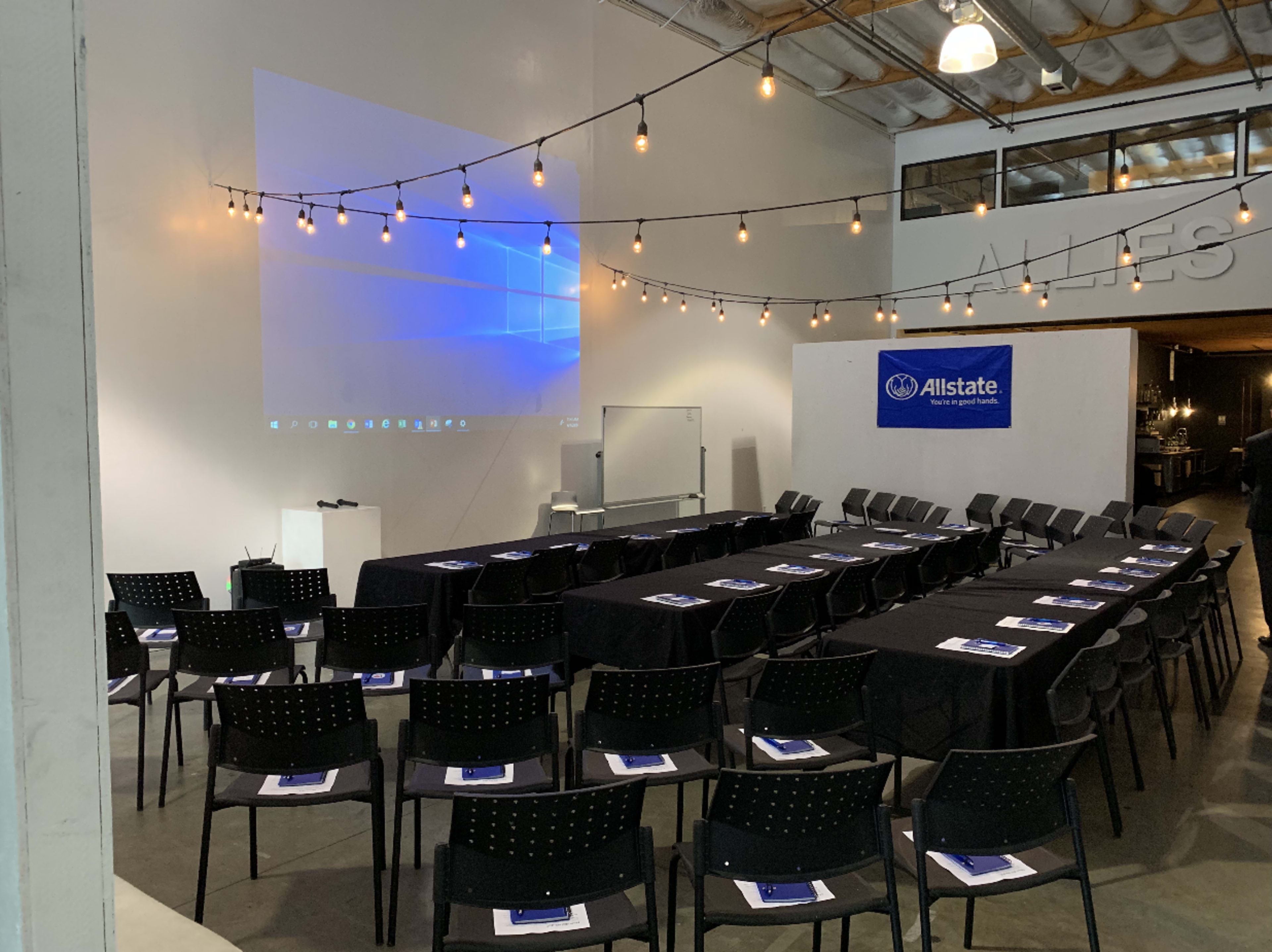 A spacious meeting room is set up with rows of chairs facing a projector screen, decorated with string lights above.