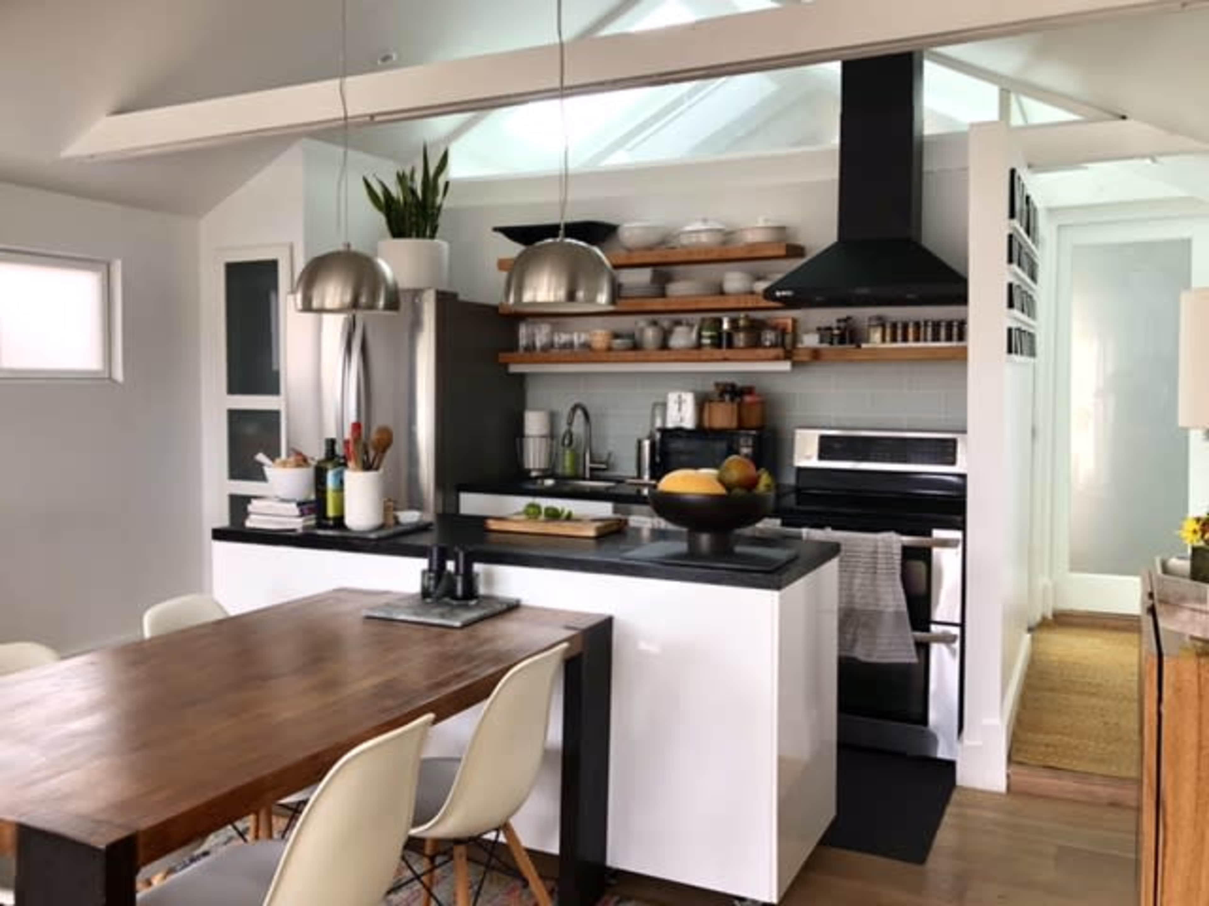 The image shows a modern kitchen featuring a central wooden dining table, sleek cabinetry, open shelving with kitchenware, and stainless steel appliances.