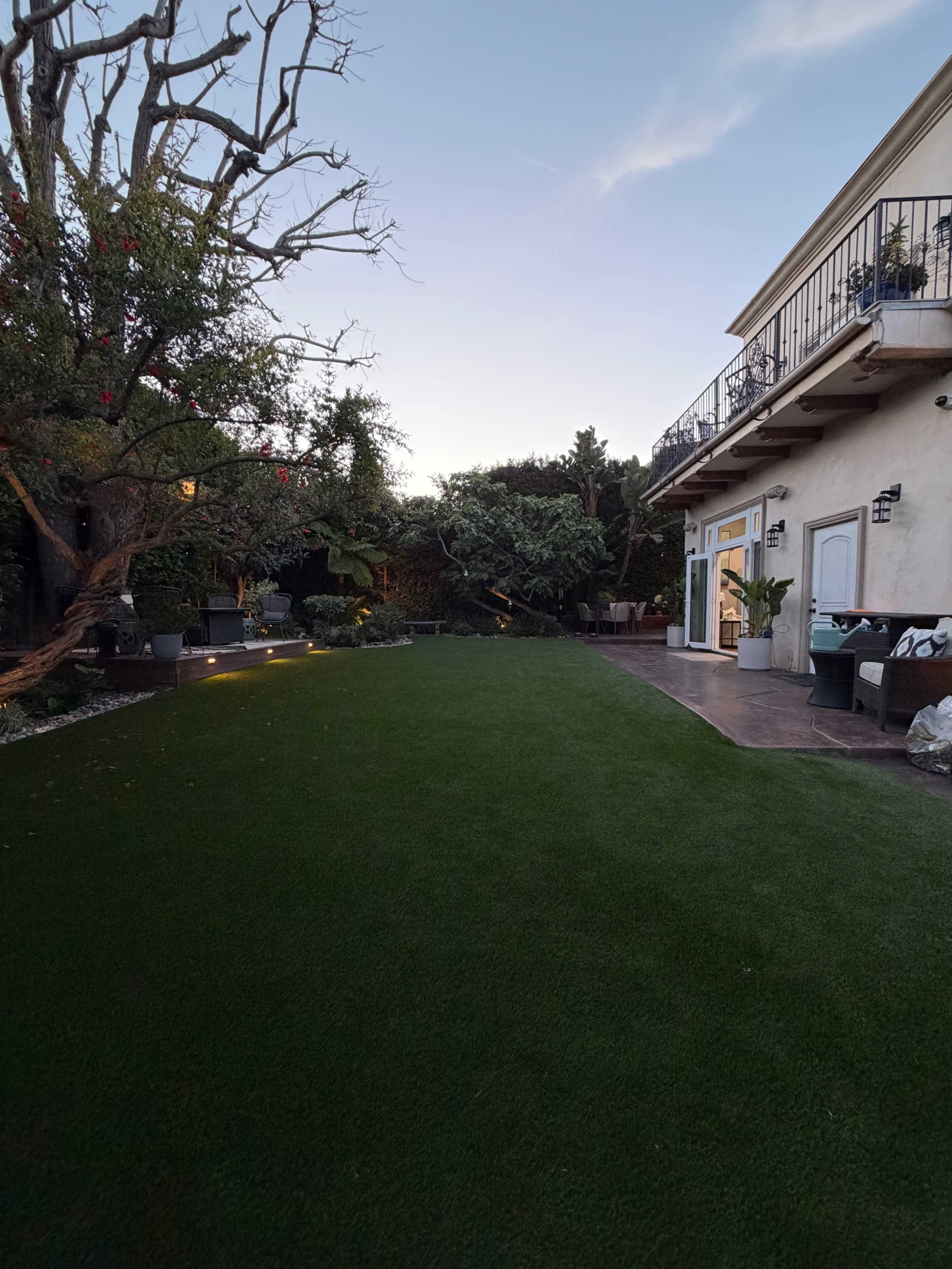 The image shows a backyard with artificial grass, trees, and a house with a balcony at dusk.