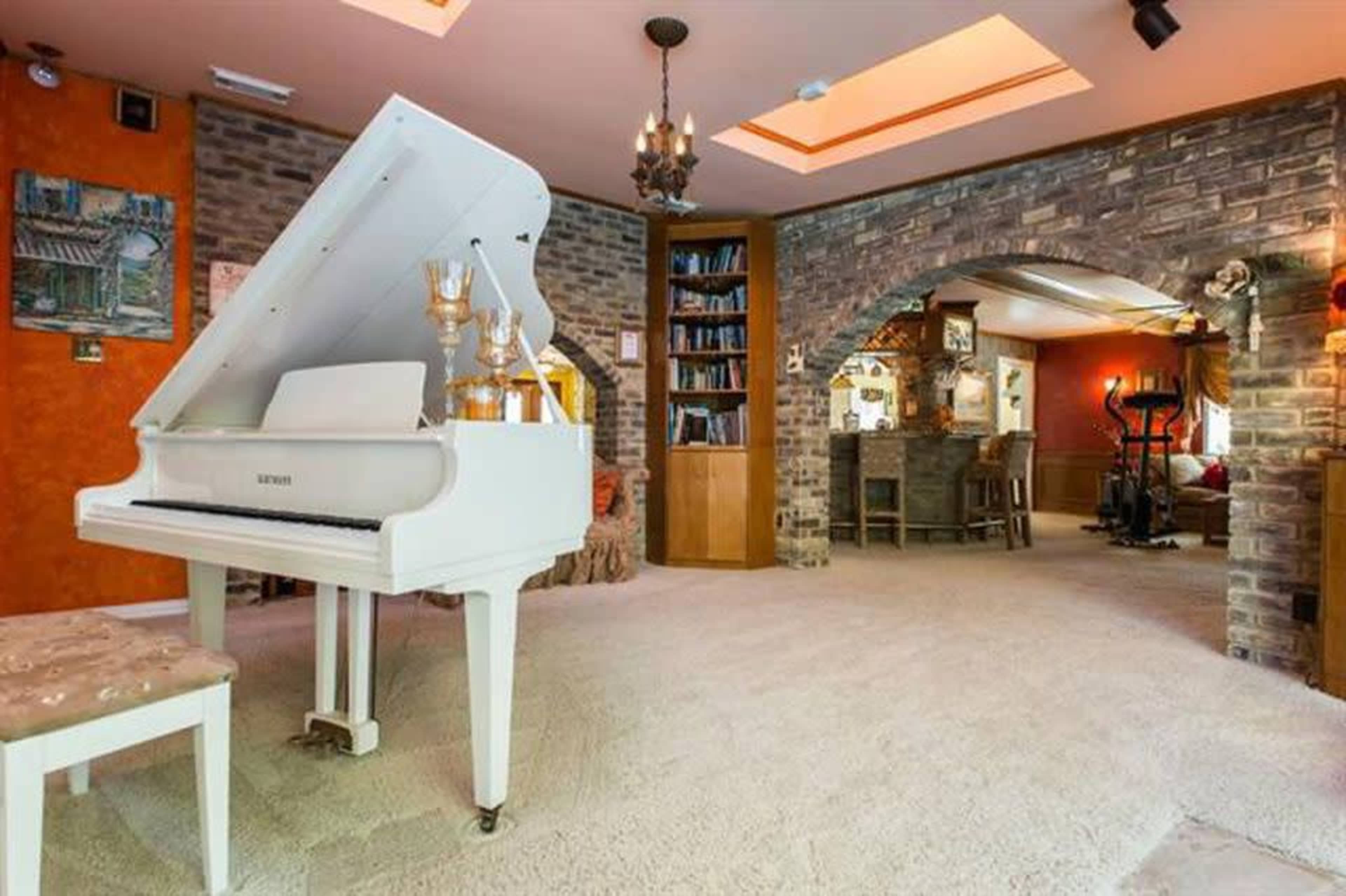 A spacious room featuring a white grand piano, a bookshelf, and various seating areas, all set against a backdrop of brick walls and warm-toned decor.