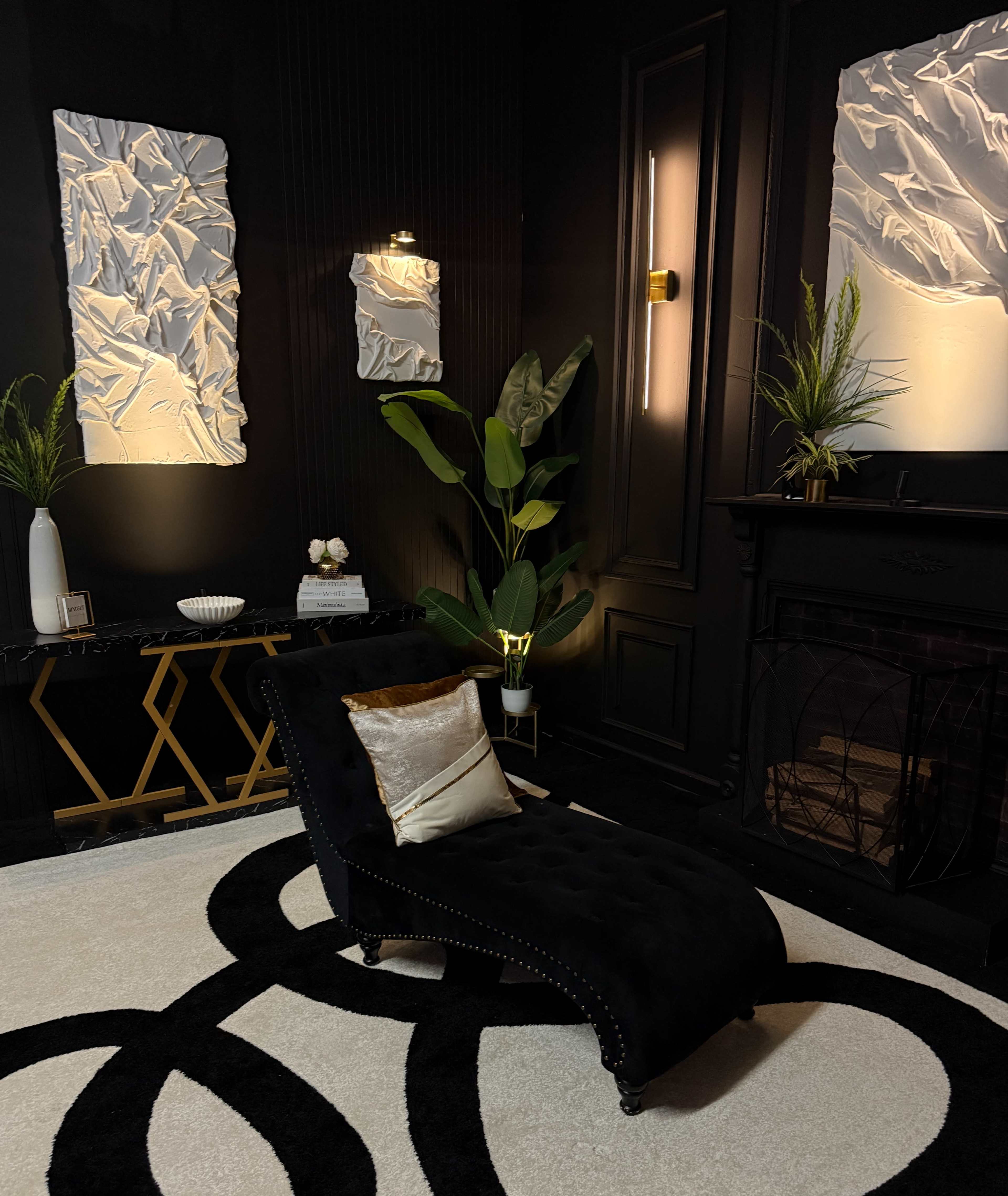 The image shows a stylish lounge area featuring a black chaise lounge, a decorative console table, plants, and textured wall panels, all against a dark wall backdrop.