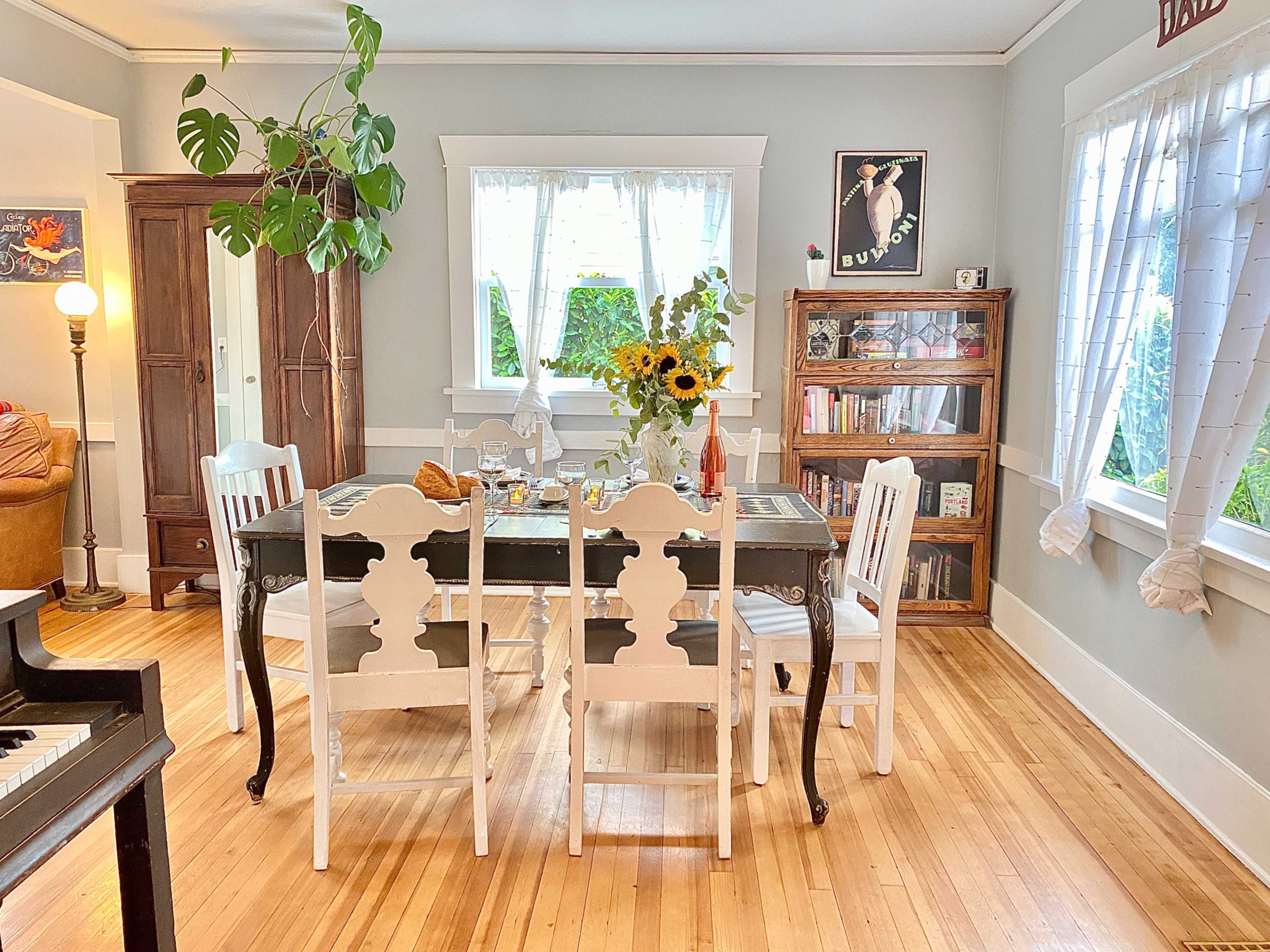 A dining area features a wooden table set with a vase of sunflowers, surrounded by white chairs, with bookshelves and a cabinet in the background.