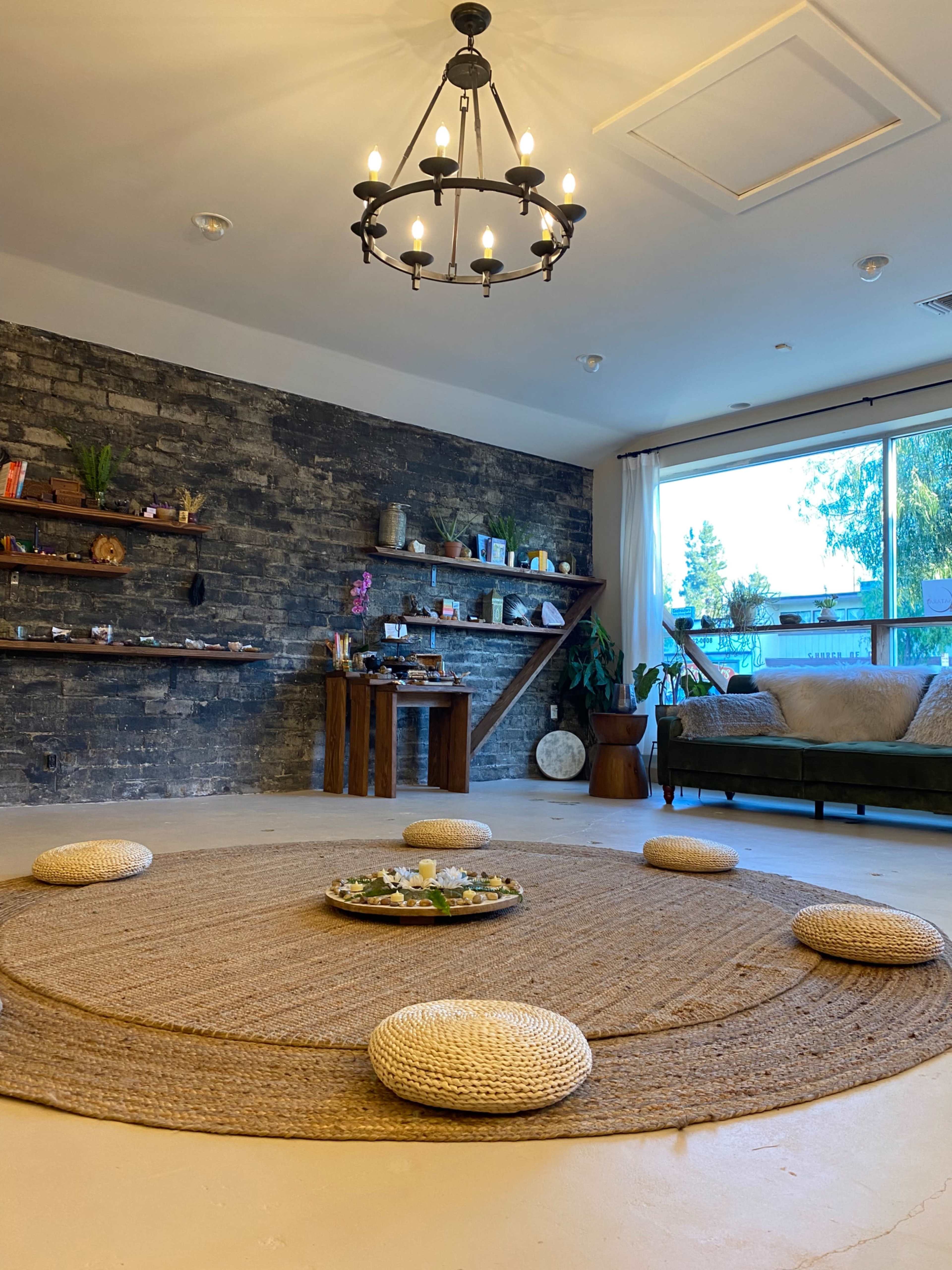 A serene interior space with a circular jute rug, floor cushions arranged around a central decorative platter, and shelves displaying various items on a textured stone wall.